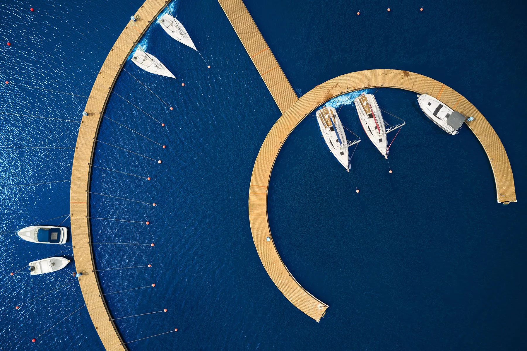 Veduta aerea di un molo con barche a vela e barche a motore ormeggiate in acque blu, con un percorso circolare in legno e boe rosse per la delimitazione dell'area nautica.