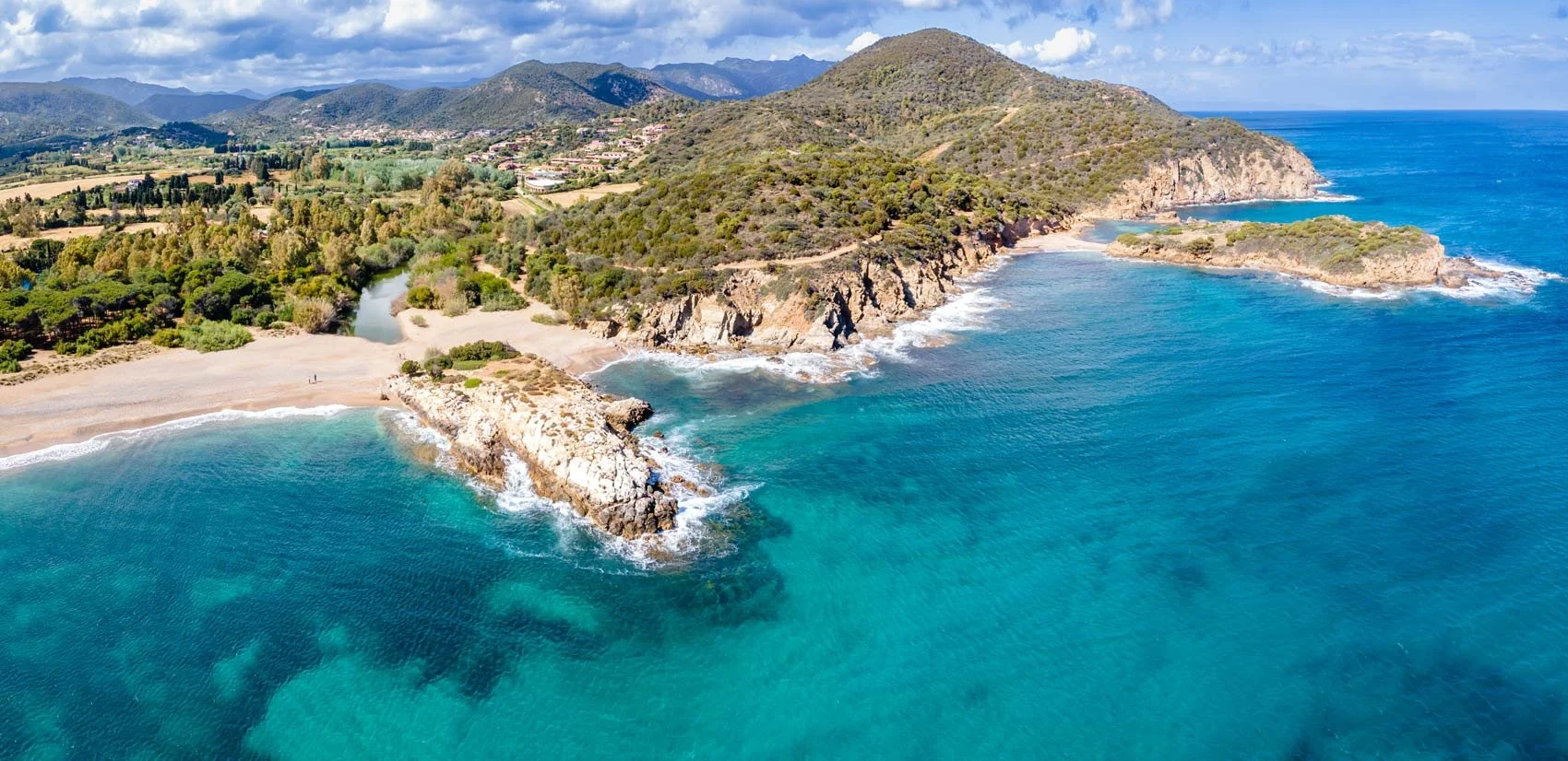 Costiera con spiaggia, scogliere e mare blu, con colline verdi e montagne sullo sfondo.