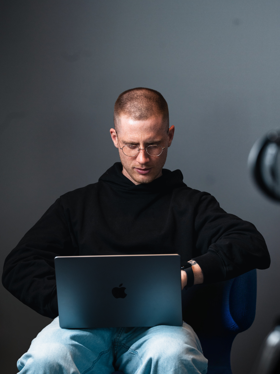 Junger Mann mit Brille sitzt vor einem Apple MacBook und schaut auf die Uhr, dunkler Hintergrund.