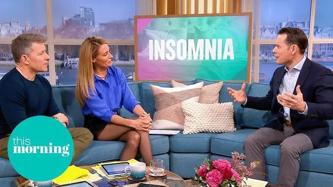 TV show set with three people in conversation. A woman in a blue blouse and black skirt, and a man in black shirt and khakis sit on a teal sofa with pillow is speaking and gesturing. Behind them is a large screen that says 'INSOMNIA'. The show's logo, 'this morning', is in the bottom left corner. A coffee table with flowers, books, and drinks is in front.