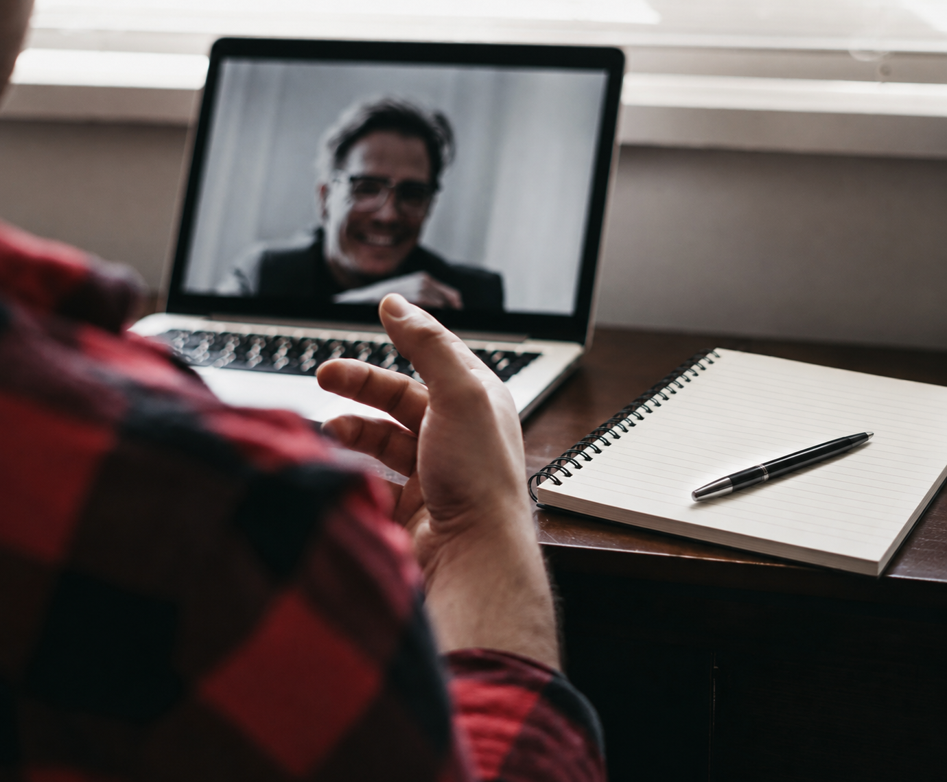 Person in red plaid shirt having a video call on a laptop, with a notebook and pen on the desk.