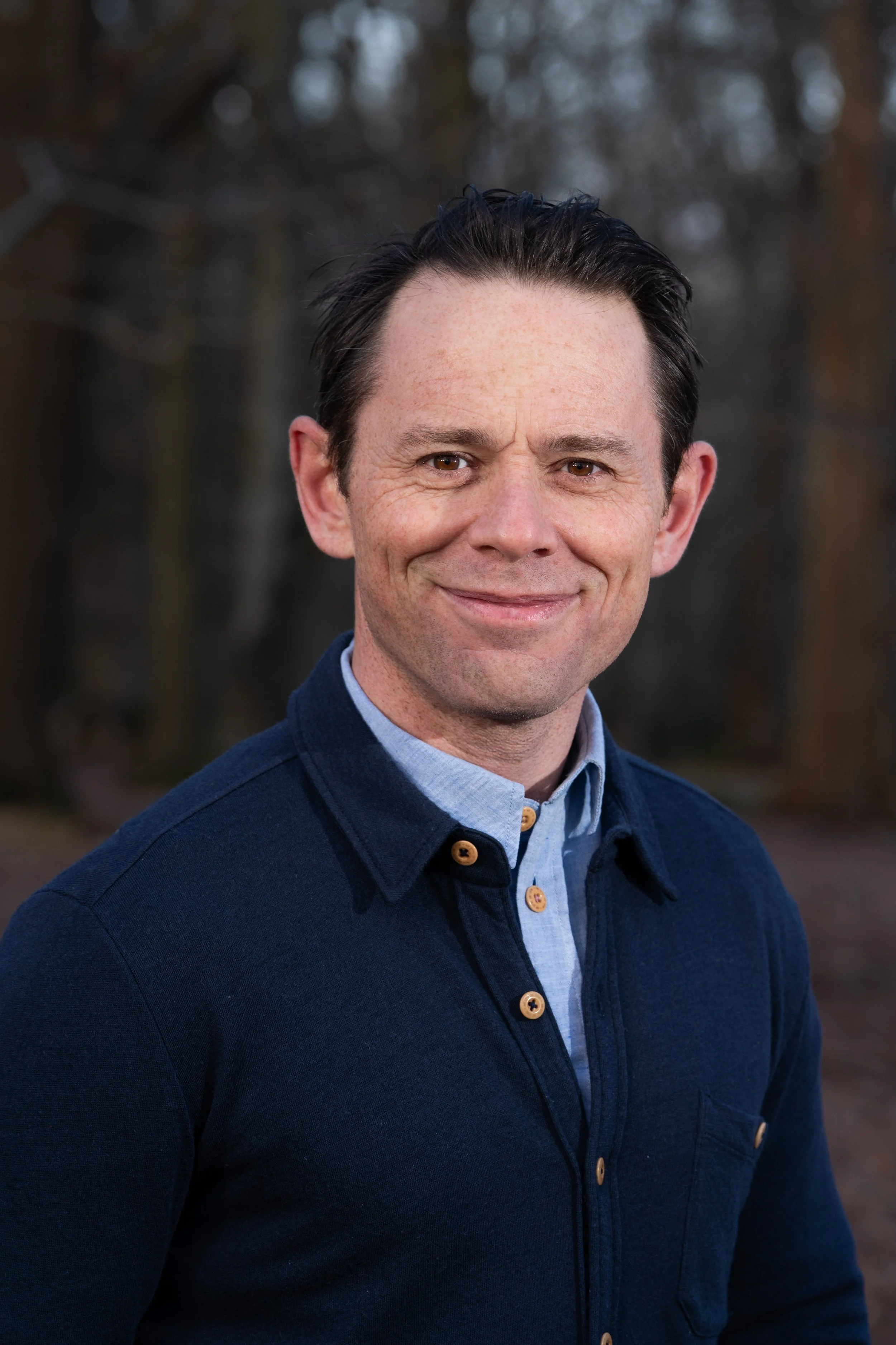 Headshot of Dr Guy Meadows with dark hair, wearing a blue cardigan over a collared shirt, outdoors with blurred trees in the background.