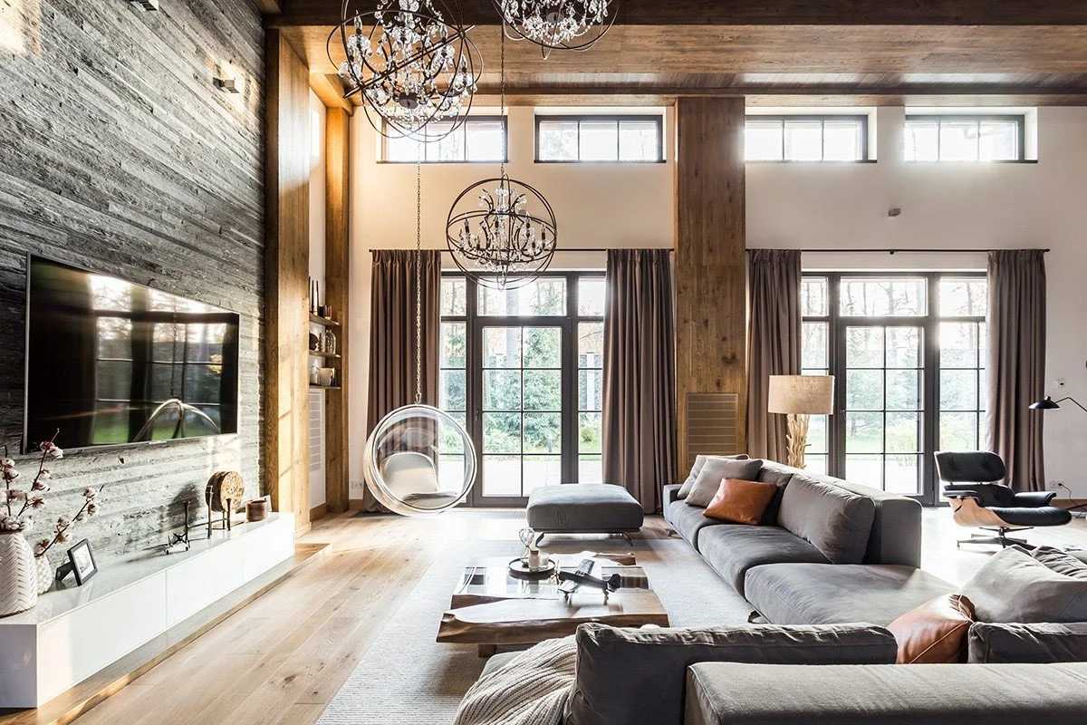 A spacious living room with large windows, beige curtains, a gray sectional sofa with pillows, a wooden floor, and modern lighting fixtures, including chandeliers and a hanging bubble chair.