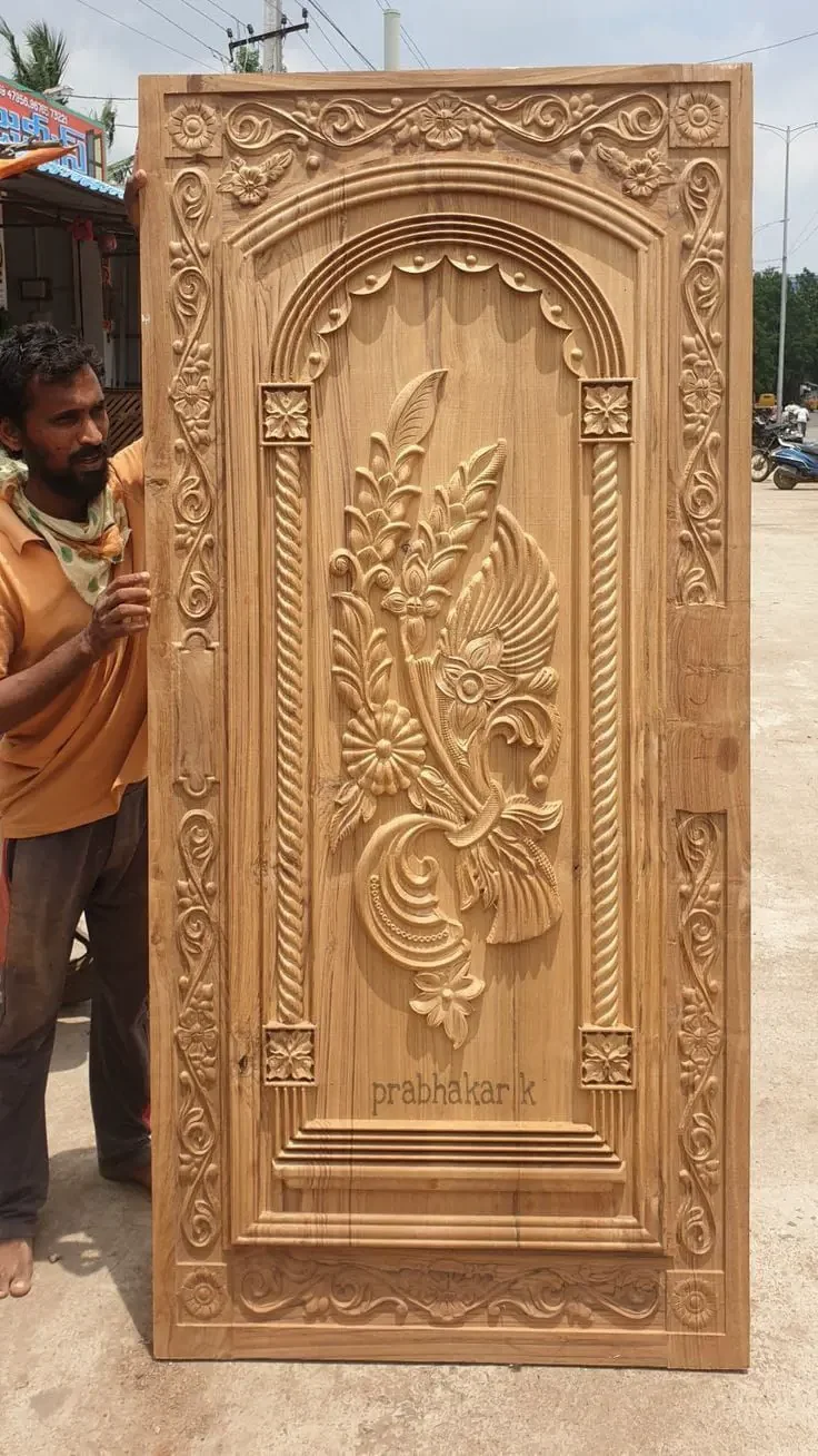 A tall, ornate wooden door with intricate carvings of a floral design and a bird, surrounded by decorative borders, and the name 'prabhakar.k' carved at the bottom. A man in a beige shirt and brown pants stands beside the door, holding it upright outdoors.