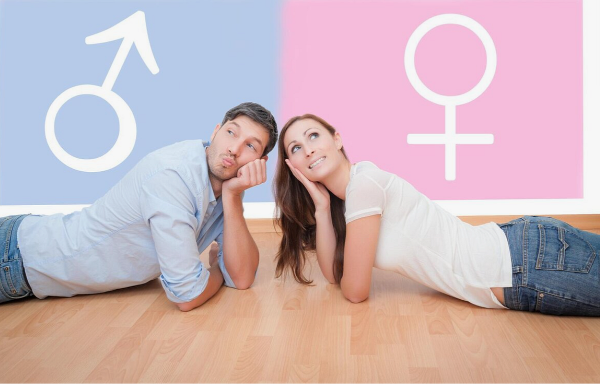 A couple lying on the floor, looking thoughtful, in front of a wall with gender symbols, a male symbol on the blue side and a female symbol on the pink side.