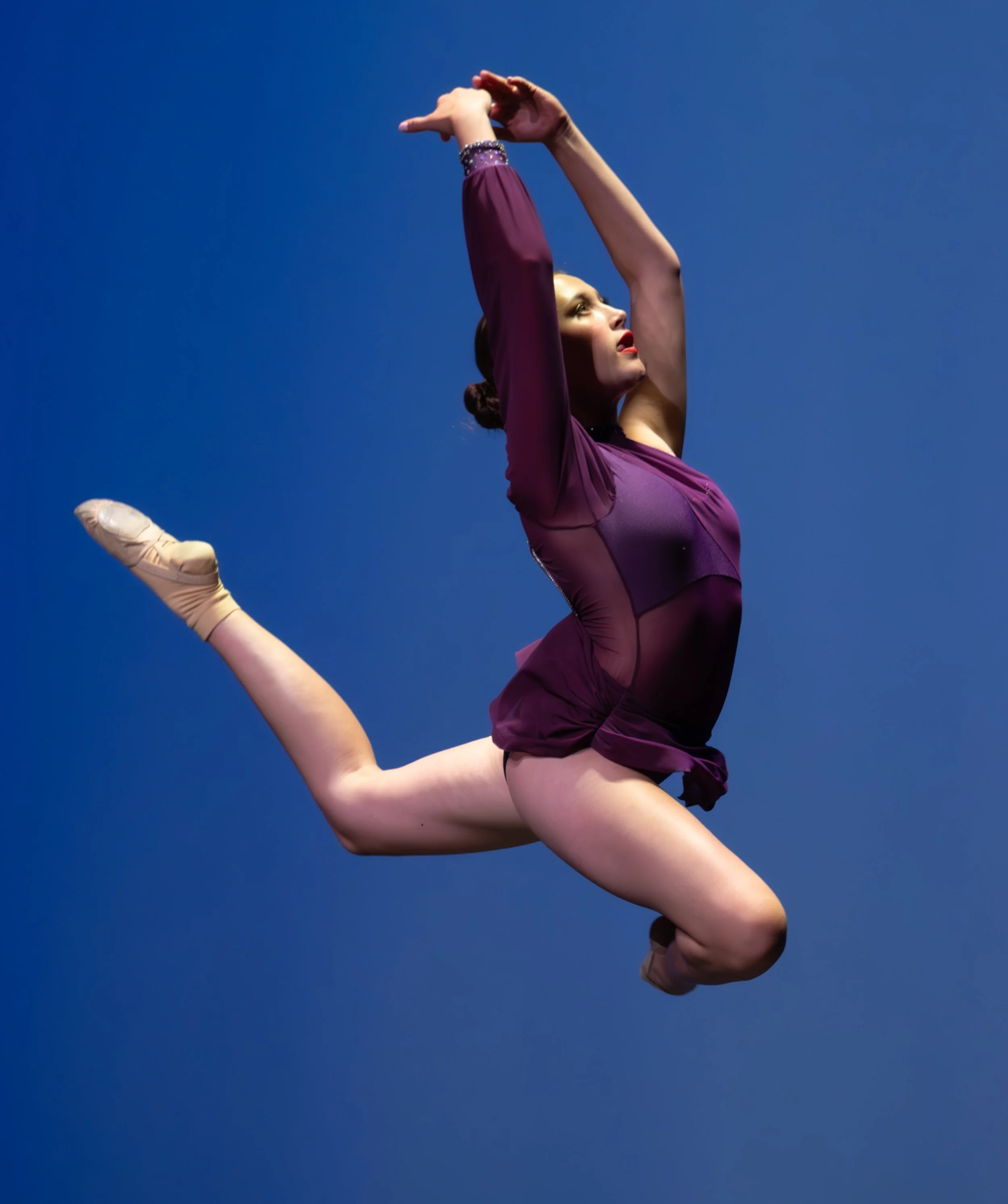 A female gymnast in a purple leotard performing a dance split mid-air against a blue background.