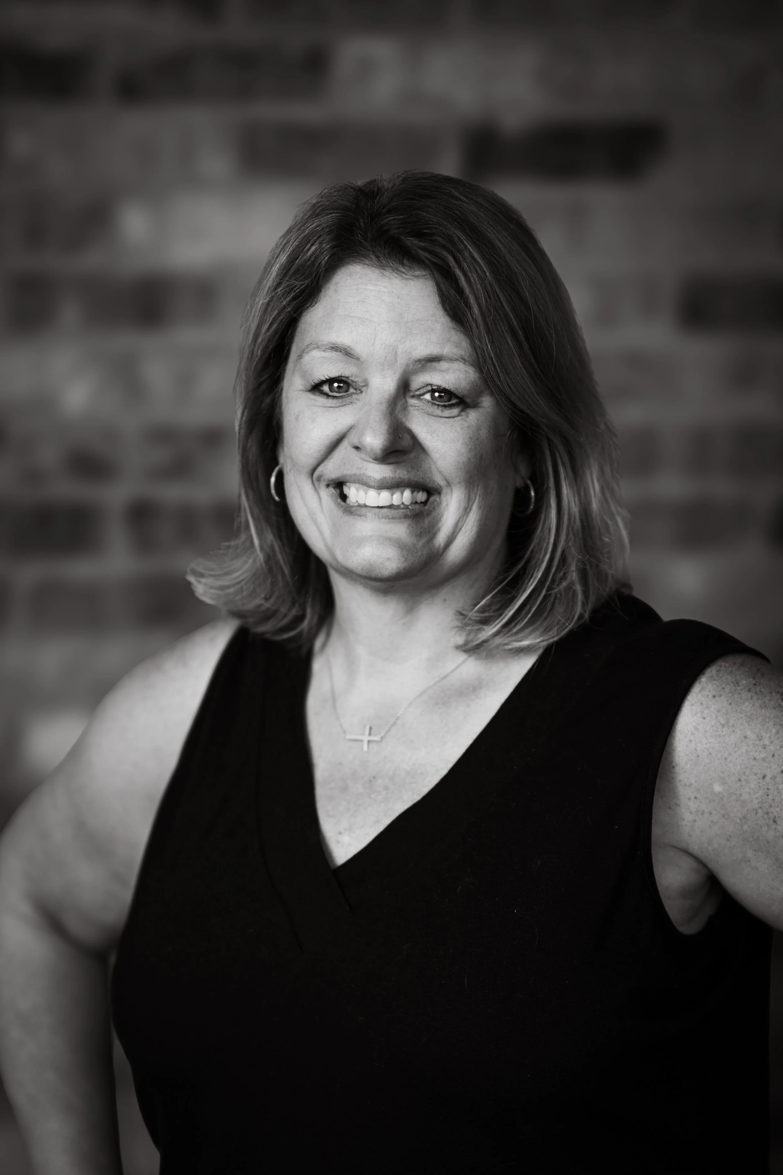 Black and white portrait of a smiling woman with shoulder-length hair, wearing hoop earrings, a cross necklace, and a sleeveless top, standing against a blurred brick wall background.