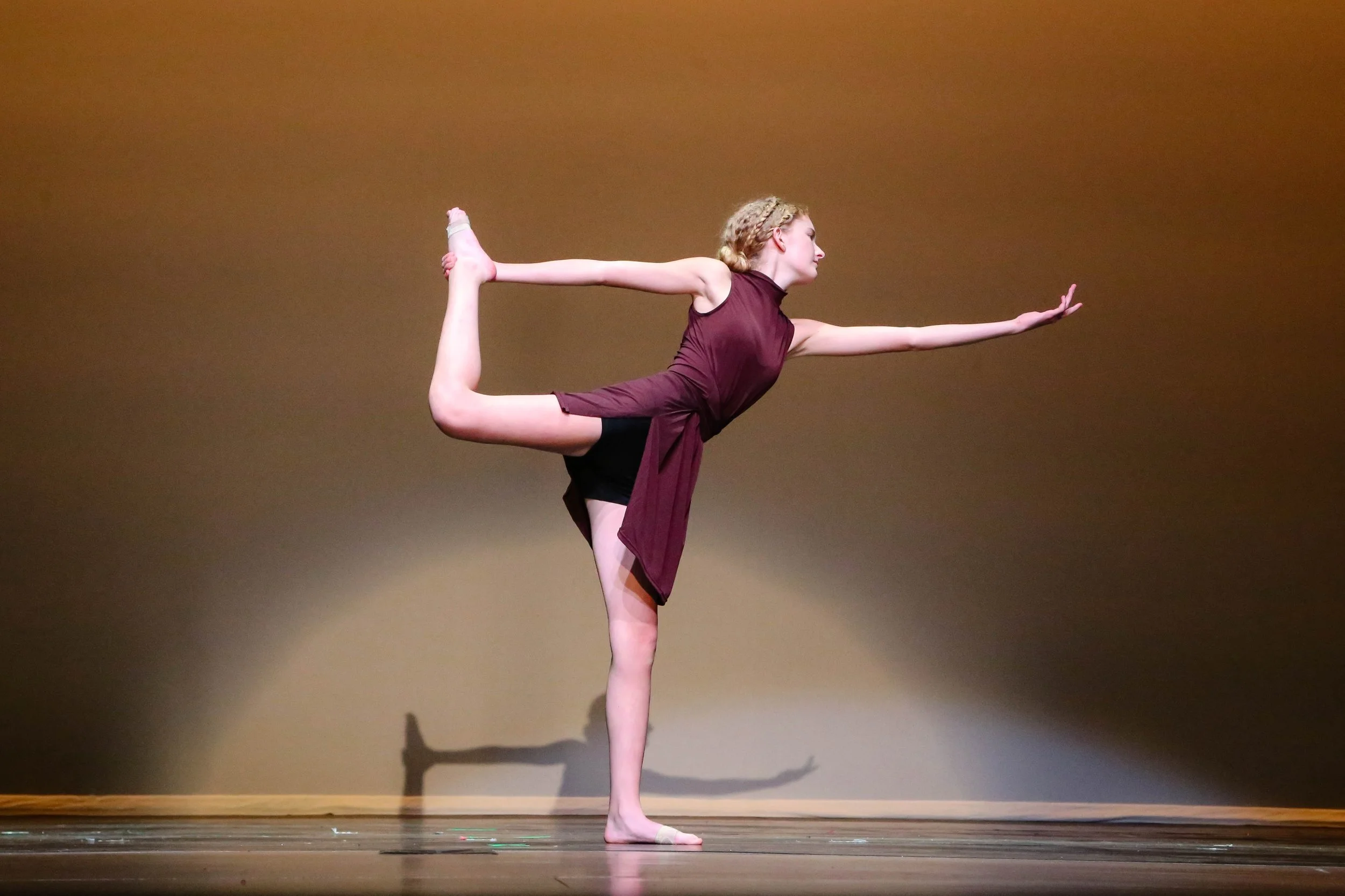A young woman performing a dance pose on stage, standing on one leg with the other bent backwards and held by her hand, while extending her other arm forward.