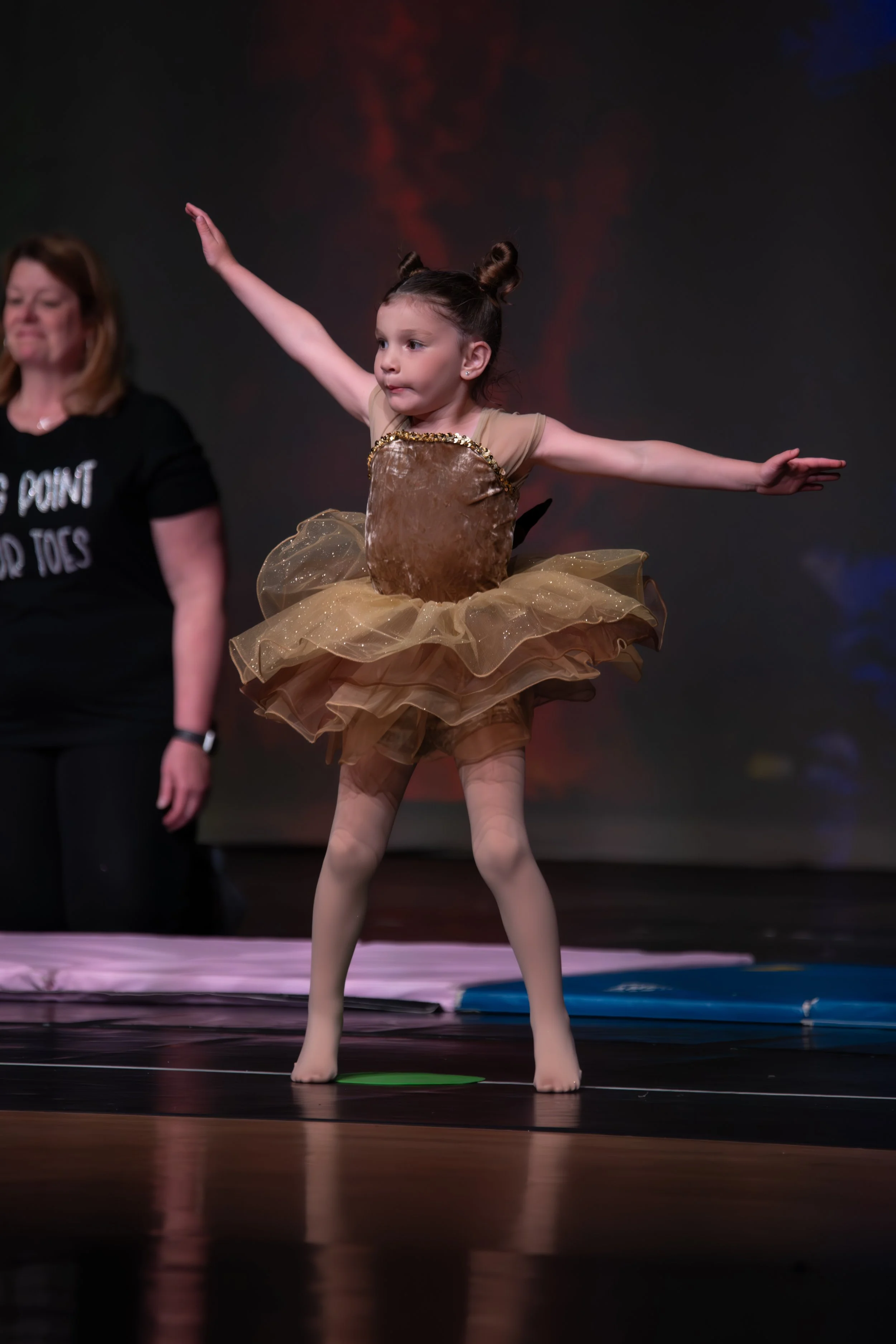 Young girl in gold ballet costume performing a ballet move on stage, with her arms extended and standing on her toes, with a woman in black shirt standing behind her.