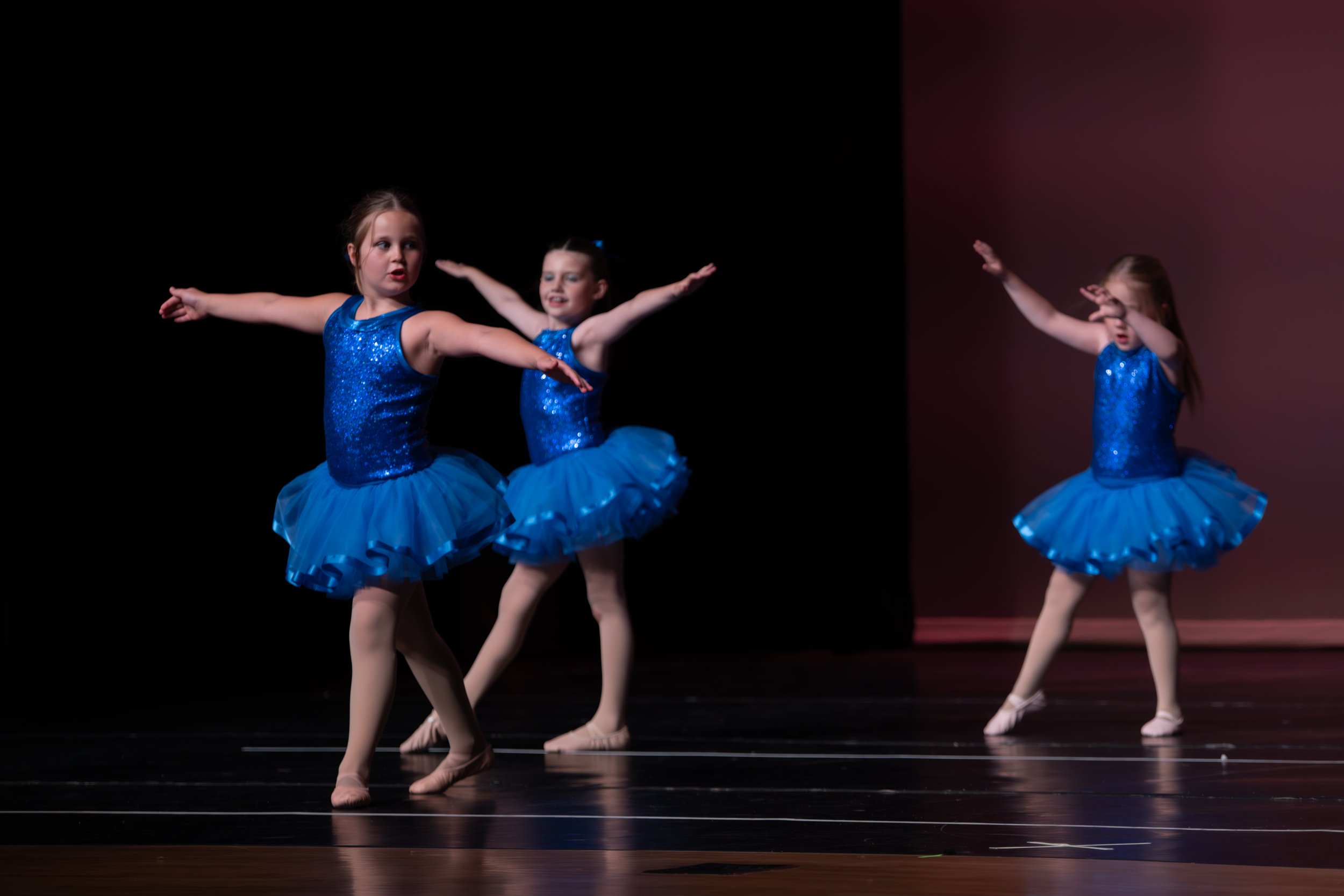 Three young girls in blue glittery ballet costumes practicing dance moves on stage with a dark background.