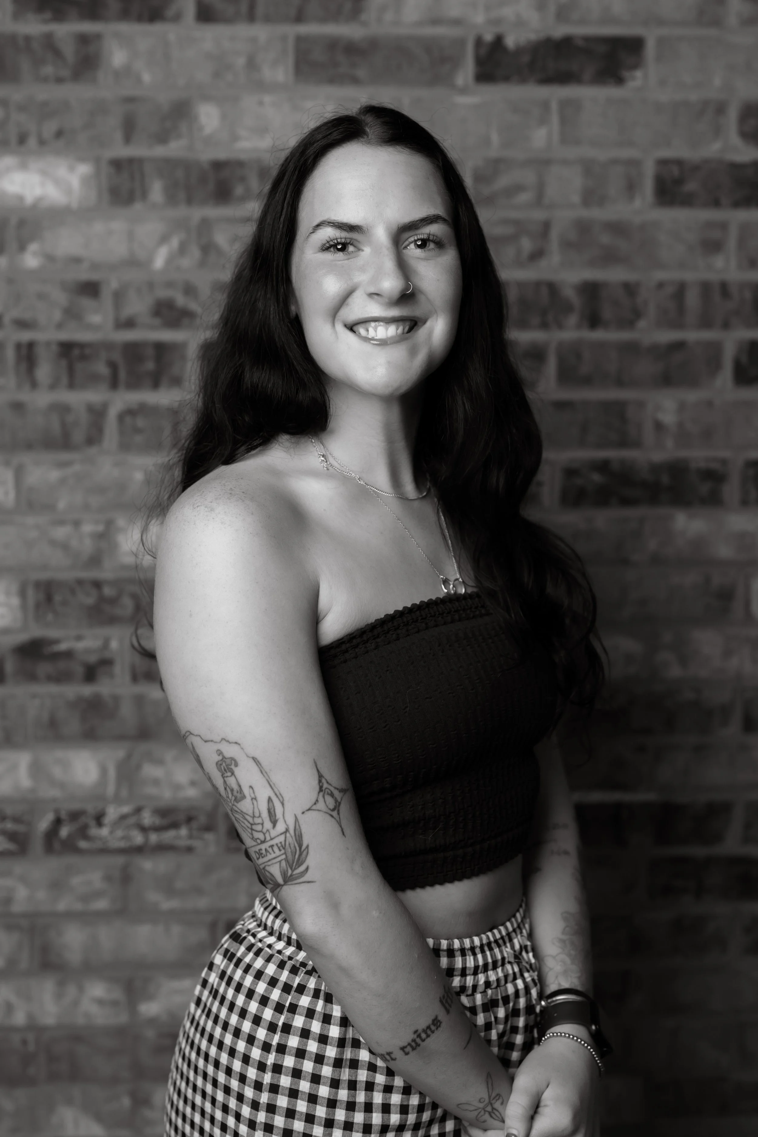 Black and white portrait of a young woman with long dark hair, smiling, standing against a brick wall. She has a nose ring, tattoos on her arms, and is wearing a strapless top, checkered skirt, and jewelry.