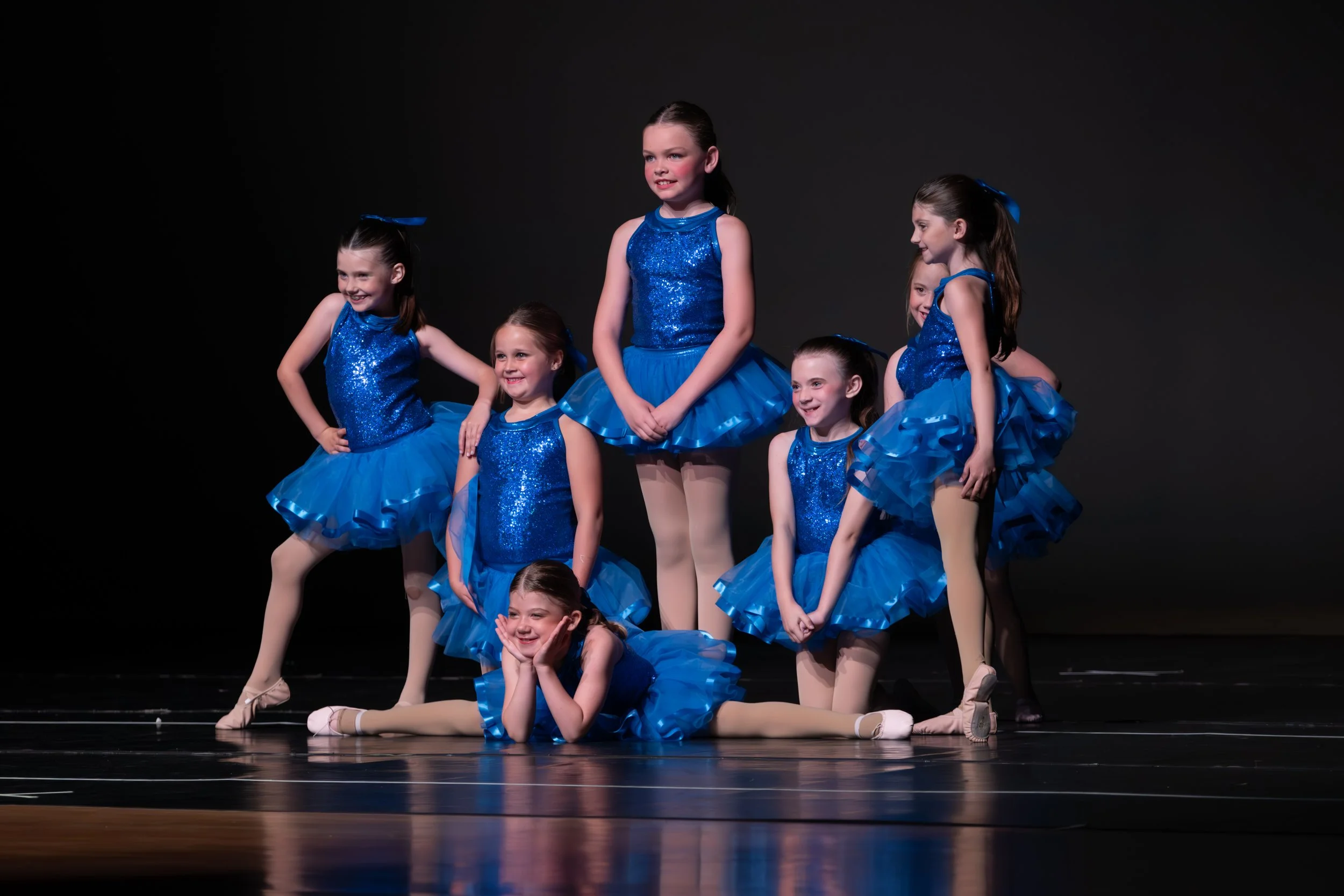 Group of young girls in blue sparkly costumes performing a ballet routine on stage with a dark background.