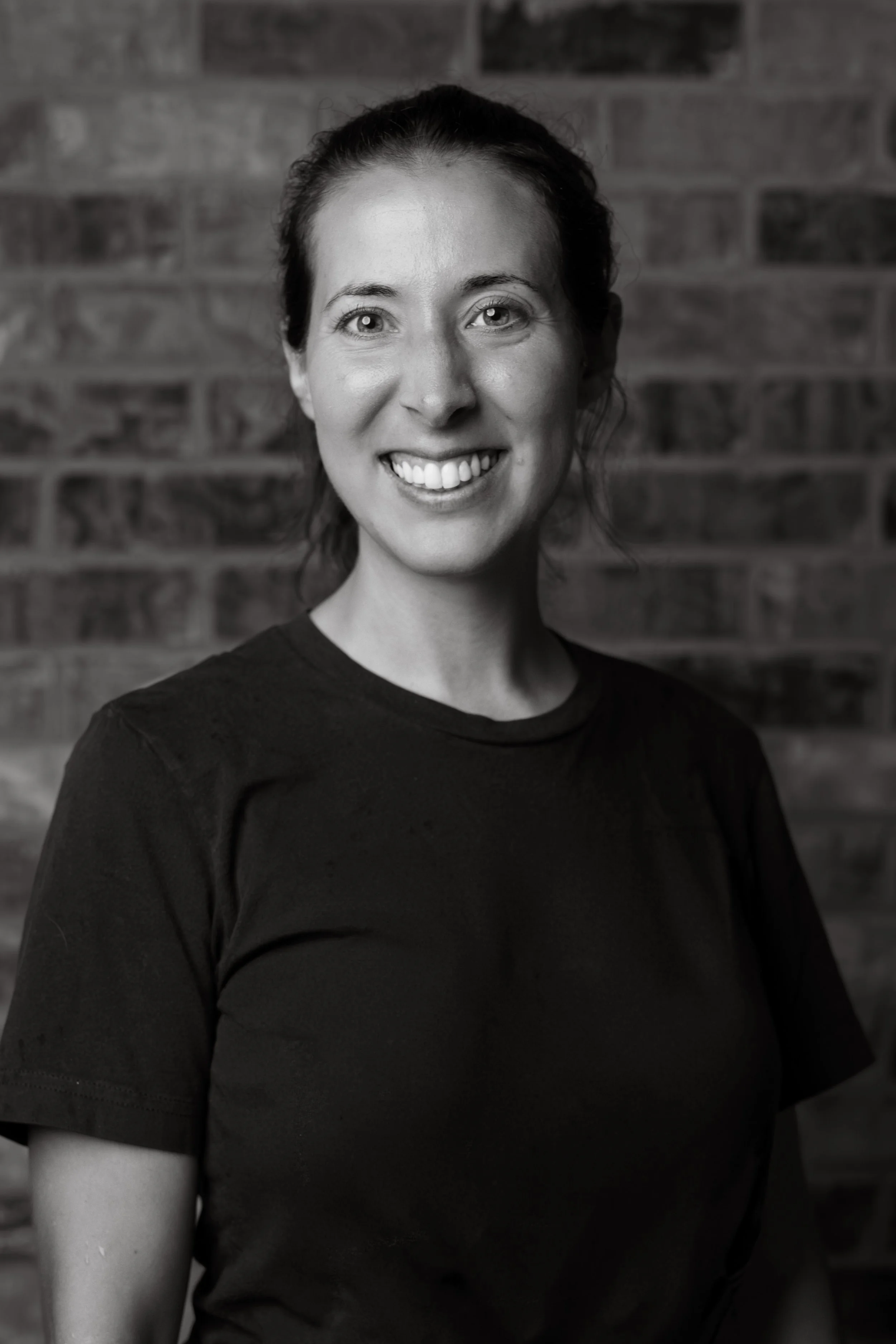 Black and white portrait of a woman with dark hair tied back, smiling, with a brick wall in the background.