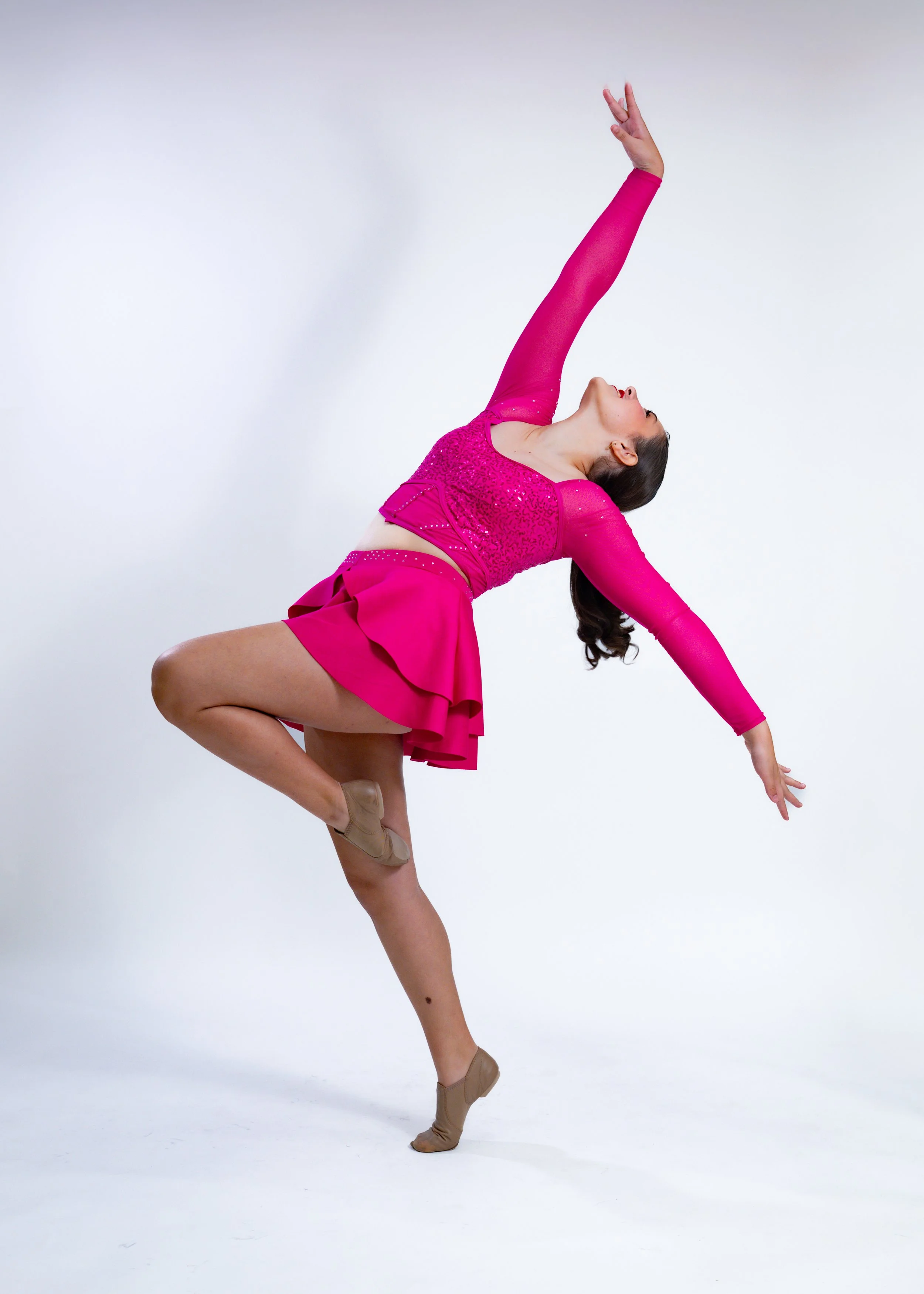 A woman in a bright pink dance costume is performing a dance pose against a plain white background. She is balancing on one leg with her other knee raised and foot pointed, while her upper body is arched backward. Her left arm is extended downward wi