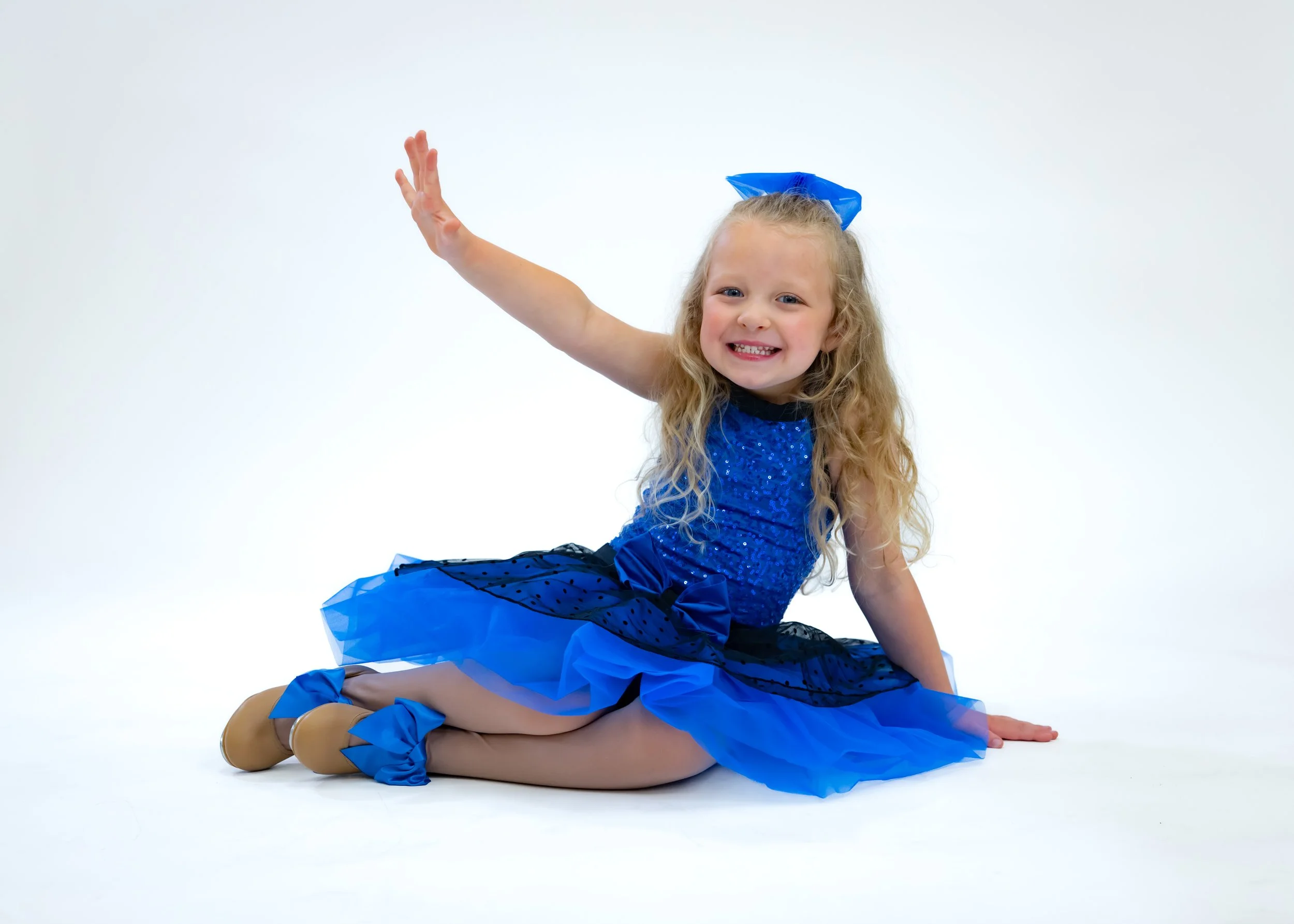 A young girl with long curly blonde hair, wearing a blue dress with sequins, tan dance shoes, and a large blue bow in her hair, is sitting on the floor with her legs folded underneath her and one arm raised, smiling at the camera against a plain whit