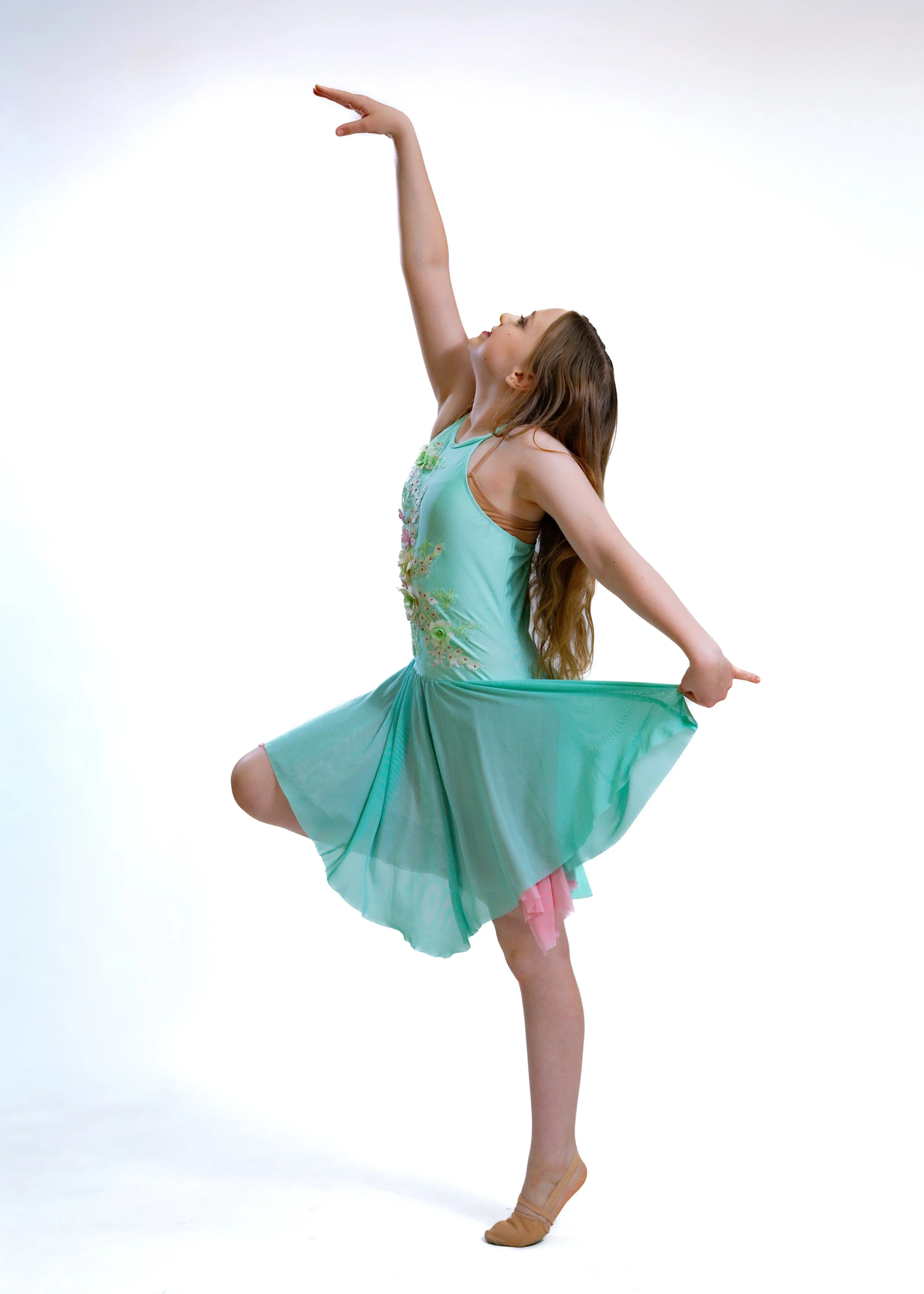 A young girl in a turquoise dress performing a dance pose on one leg, with her arms raised and head tilted back, against a plain white background.