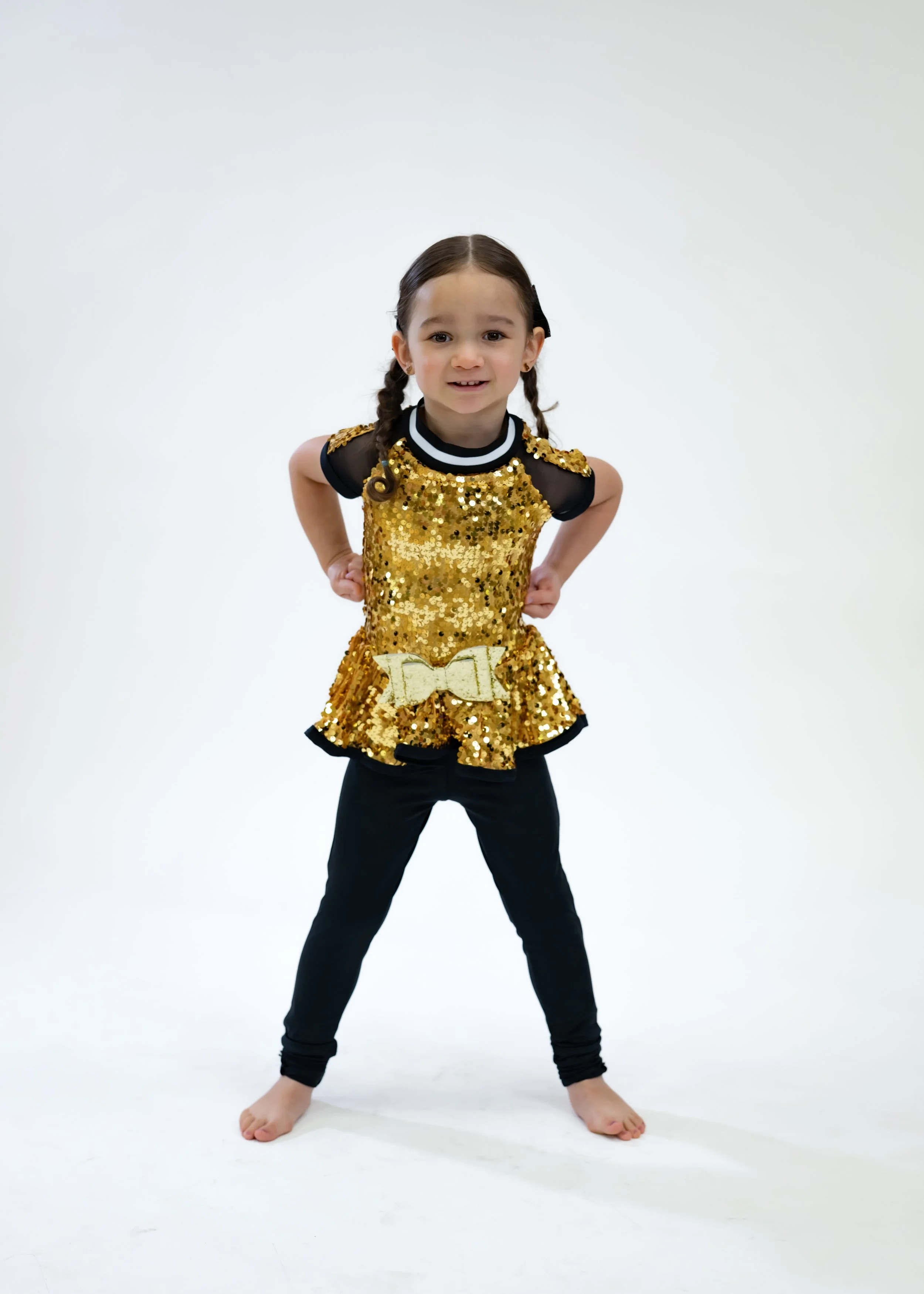 Young girl in a gold sequin top with black sleeves and pants, posing with hands on hips against a white background.