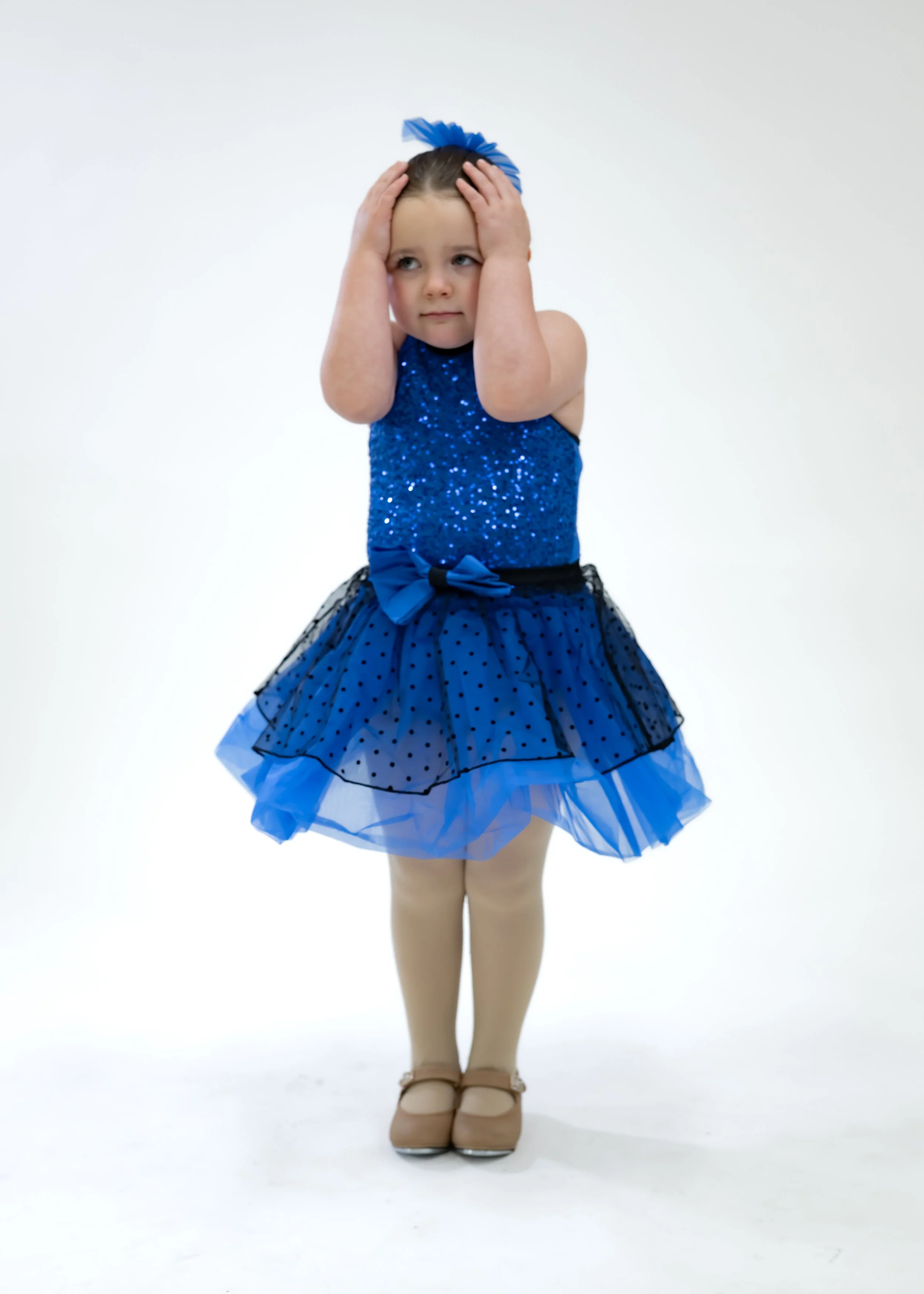 Young girl in a blue sparkly dress with a layered tulle skirt, standing on a plain white background, wearing ballet shoes.