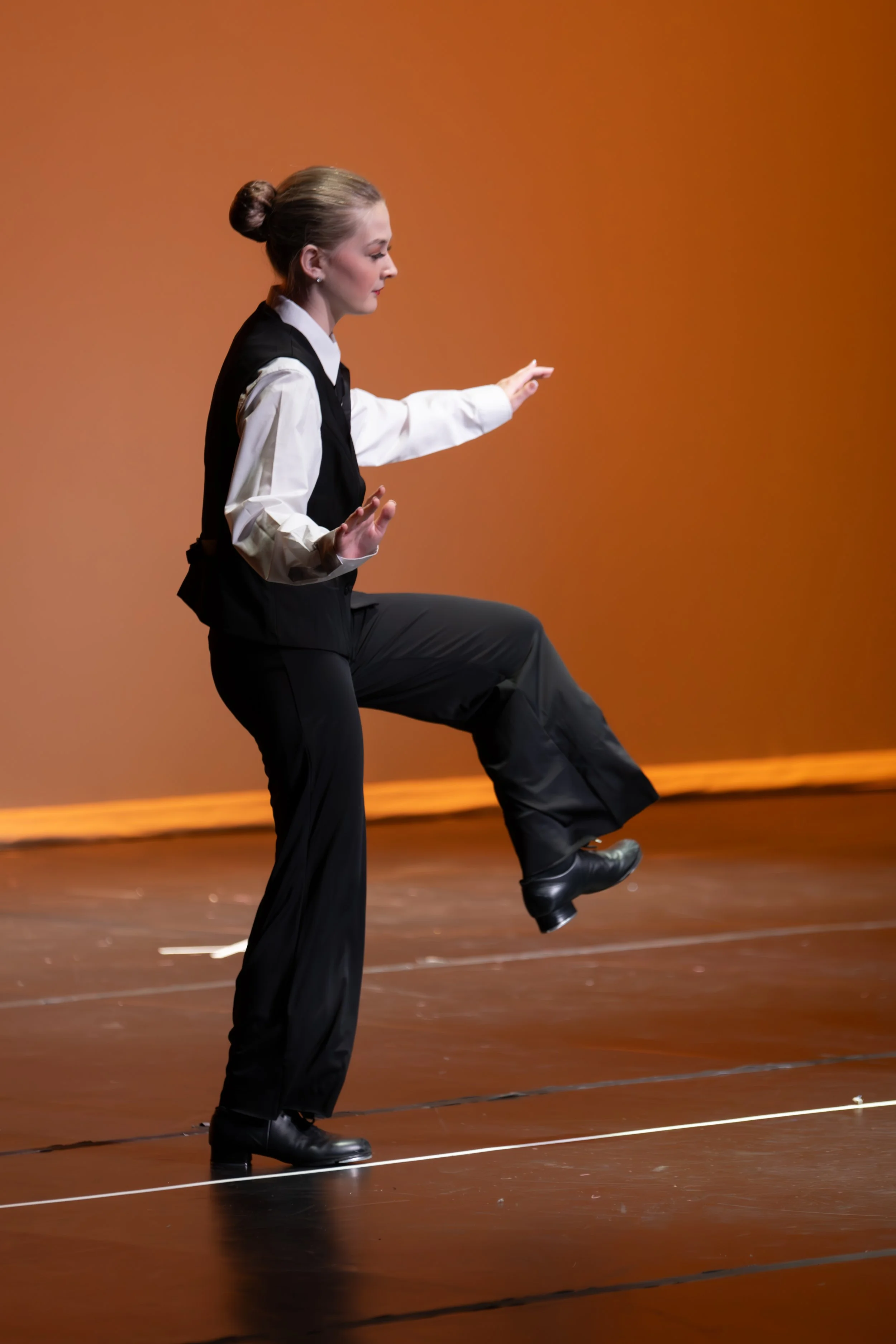 A woman in formal attire, with her hair in a bun, dance pose on stage with one leg lifted and arms extended. The stage has a plain orange background.