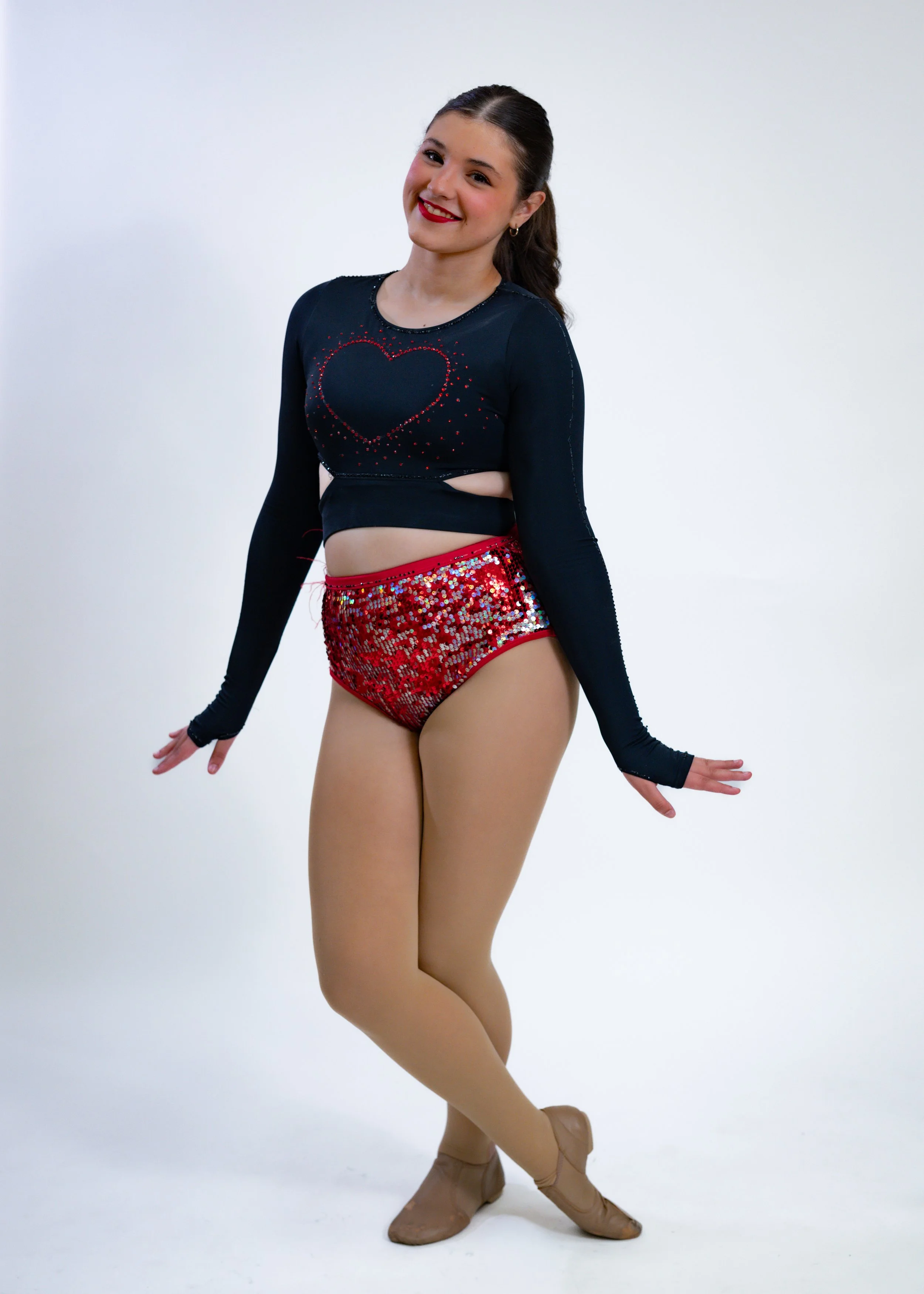 A young female gymnast in a black long-sleeve top with a heart design and red sequin shorts, posing with one leg crossed in front of the other and arms extended, wearing tan tights and beige ballet shoes, standing on a plain white background.