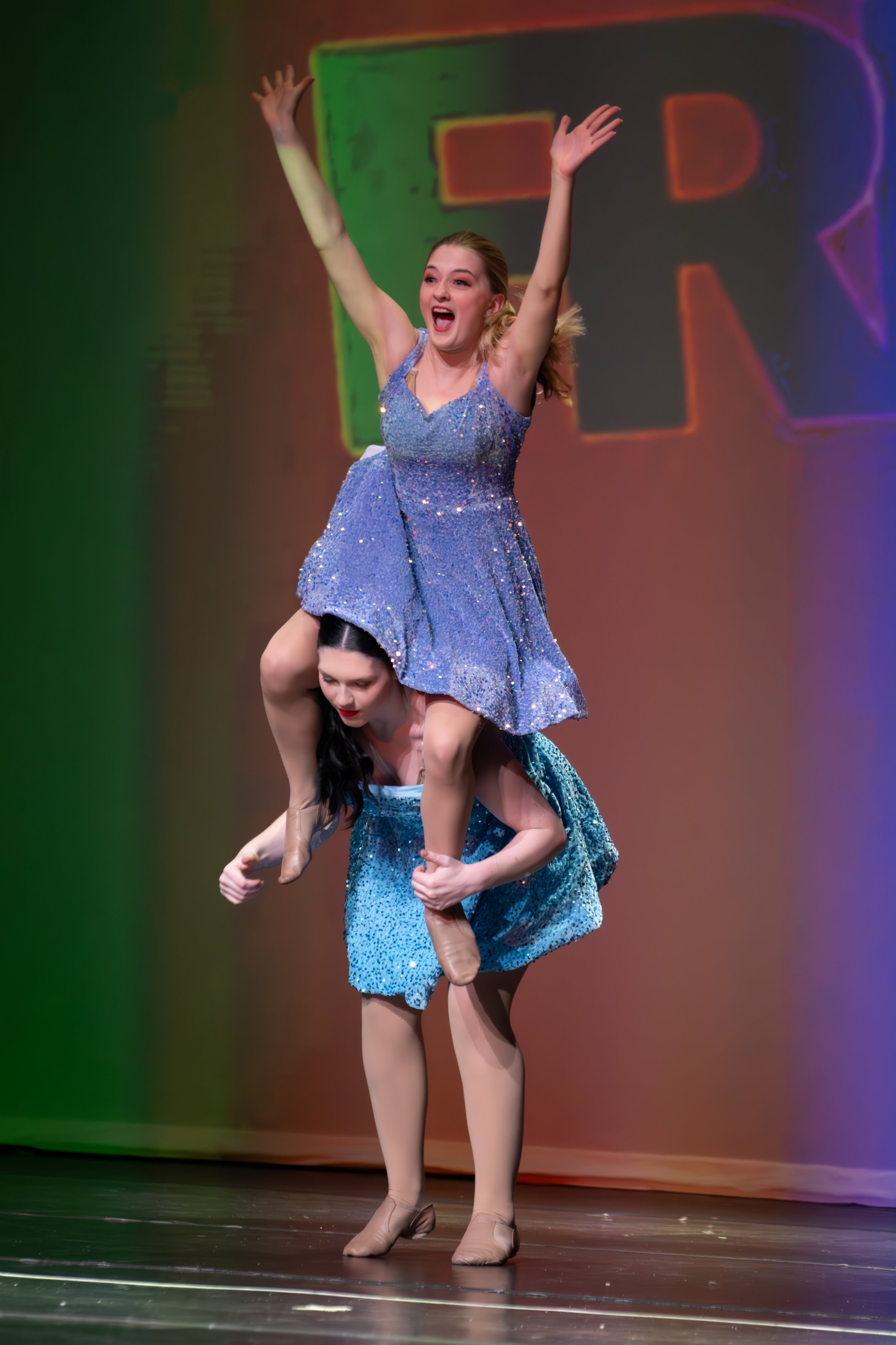 Two female performers are on stage; one is standing on the other's shoulders in a dance routine. The performer on top is wearing a sparkly blue dress with outstretched arms and an excited expression. The background features colorful lighting and the 