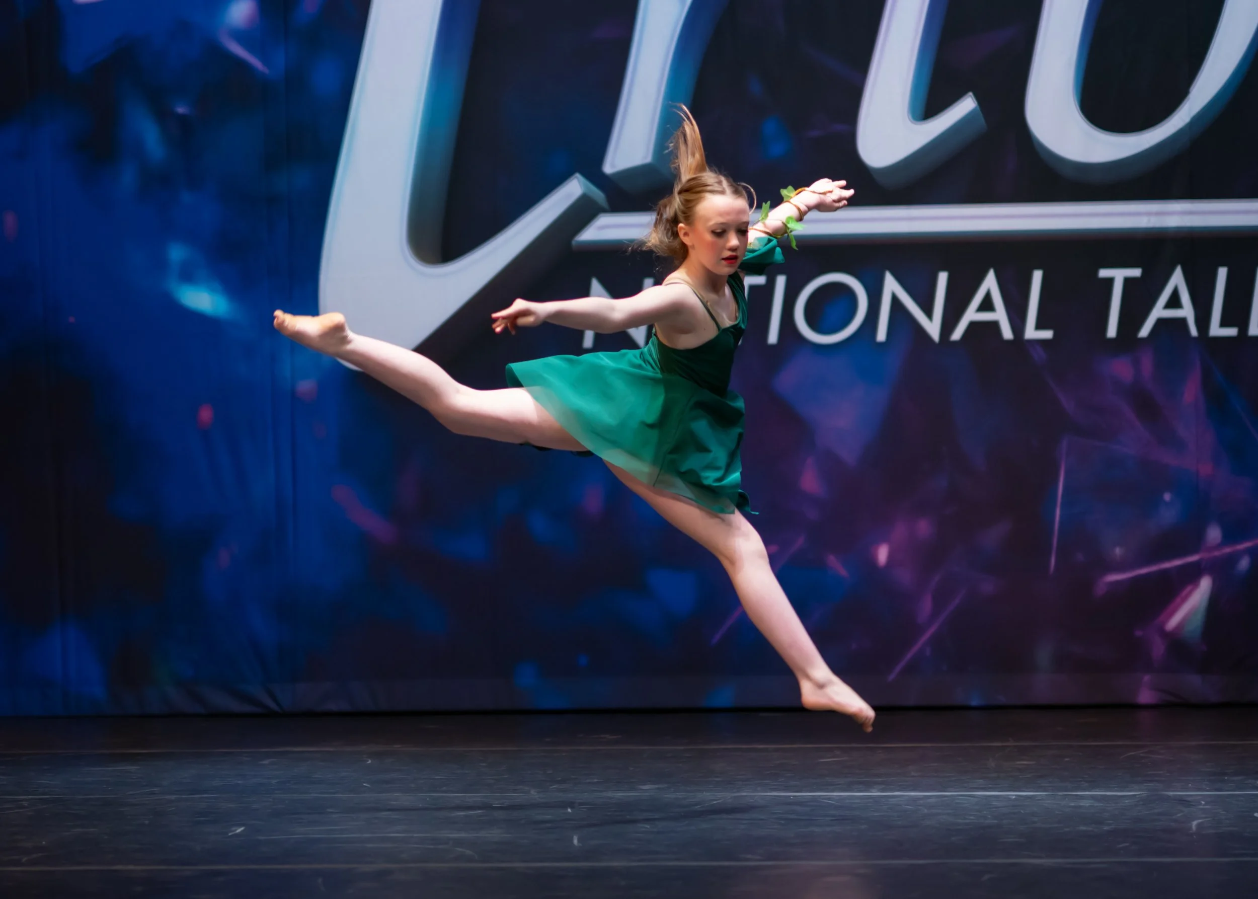 A young female dancer performing a leap on stage at a competition, with a dark backdrop displaying the word 'NATIONAL.