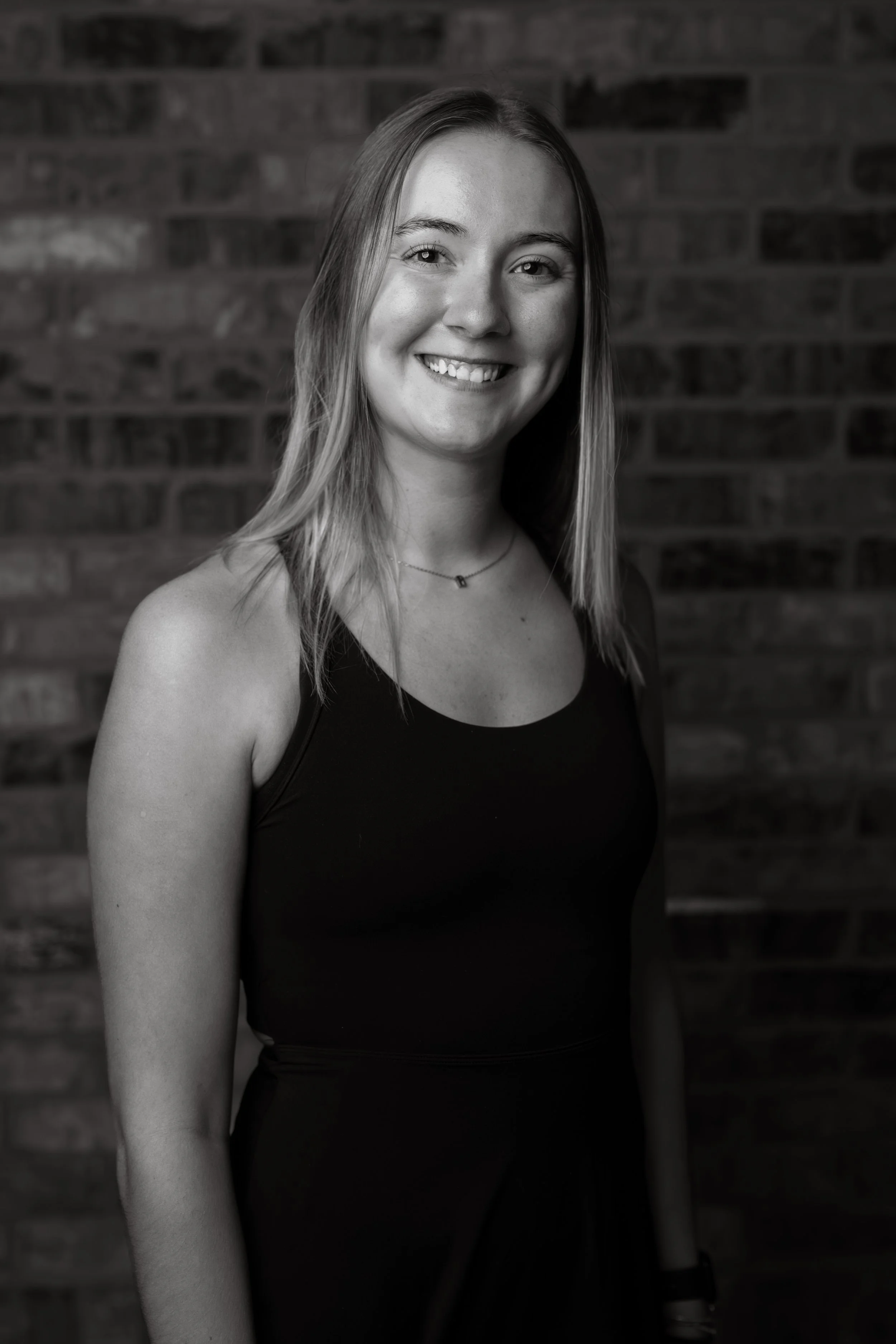 Black-and-white photo of a young woman with long hair, smiling, wearing a sleeveless top and a necklace, standing in front of a brick wall.