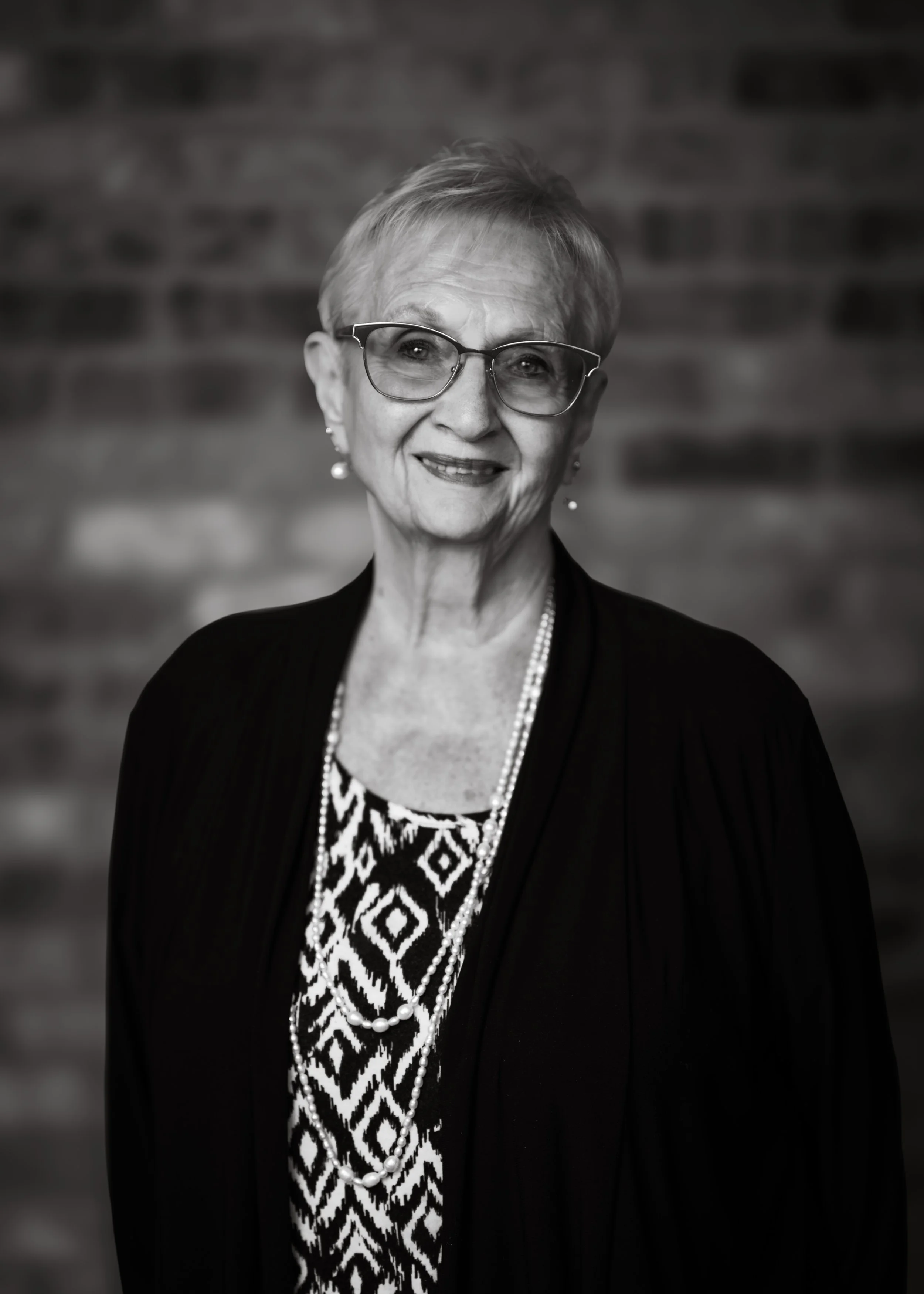 A portrait of an elderly woman with short hair, wearing glasses, a patterned top, a necklace, pearl earrings, and a dark jacket, standing in front of a brick wall.