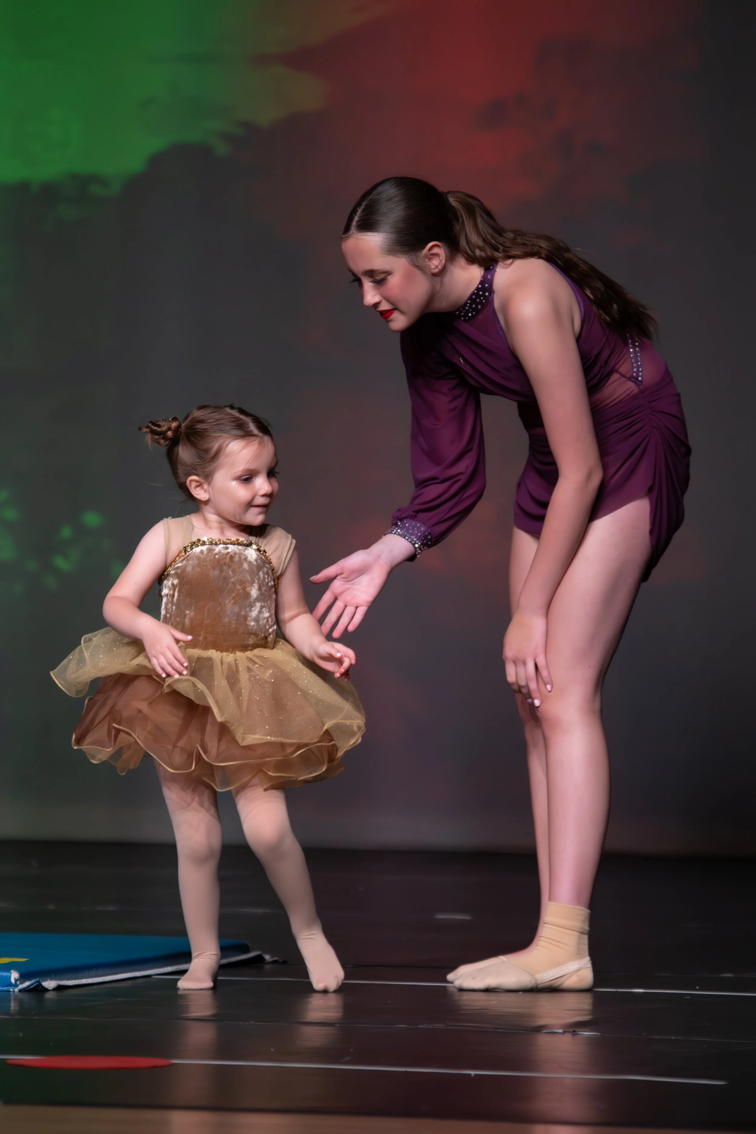 A young ballet dancer in a beige and gold tutu standing next to an older dancer in a purple costume, both wearing ballet slippers and performing on stage with a dark background.