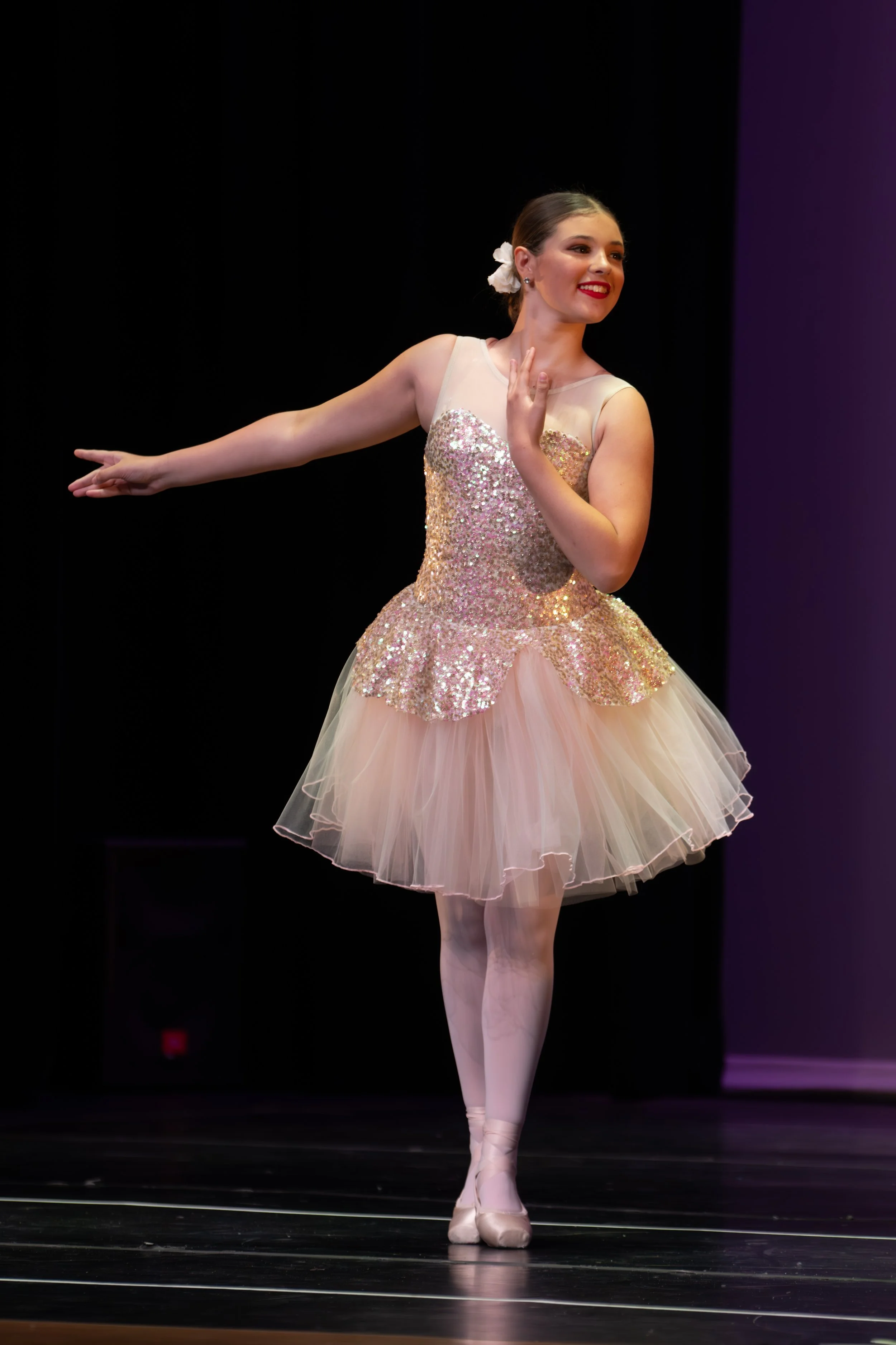 Ballet dancer in pink tutu on stage, smiling, arm extended, with dark background.