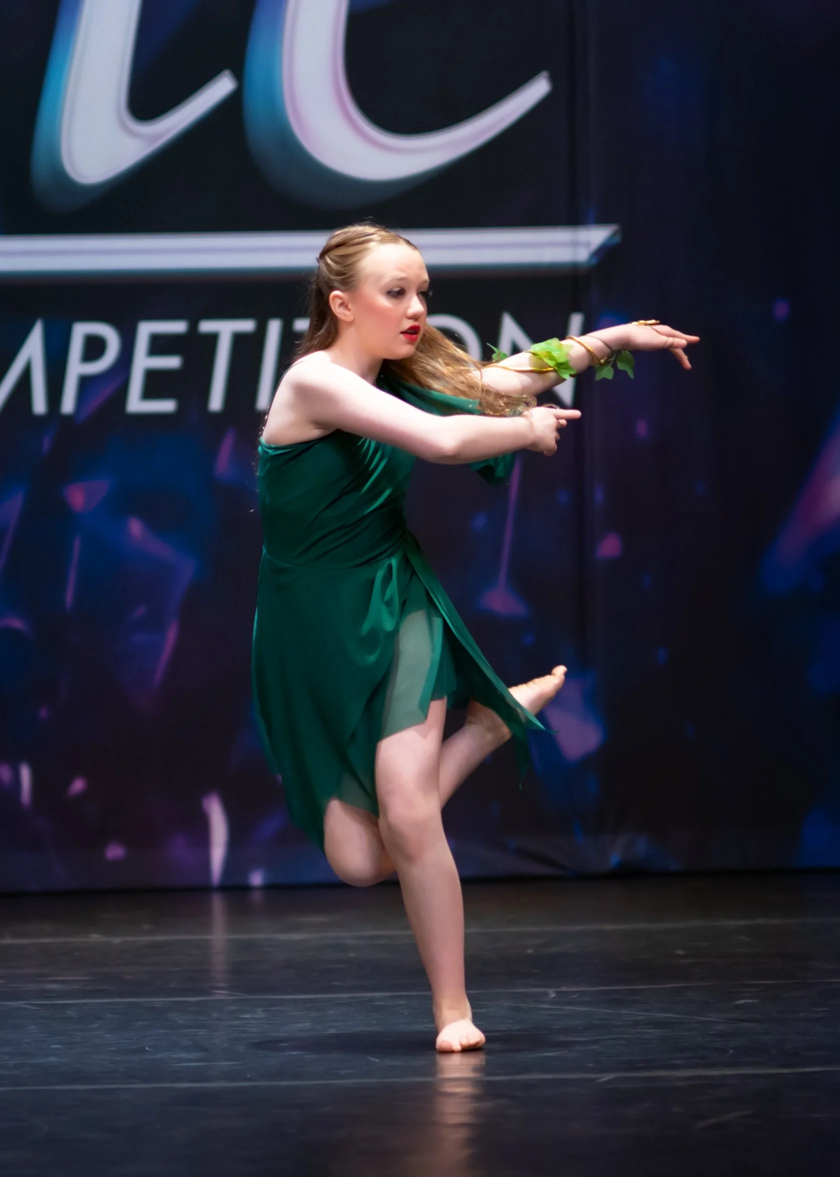 A female dancer in a green dress performing a dance pose on stage, with her arms extended and one leg lifted, against a dark background with neon lights.