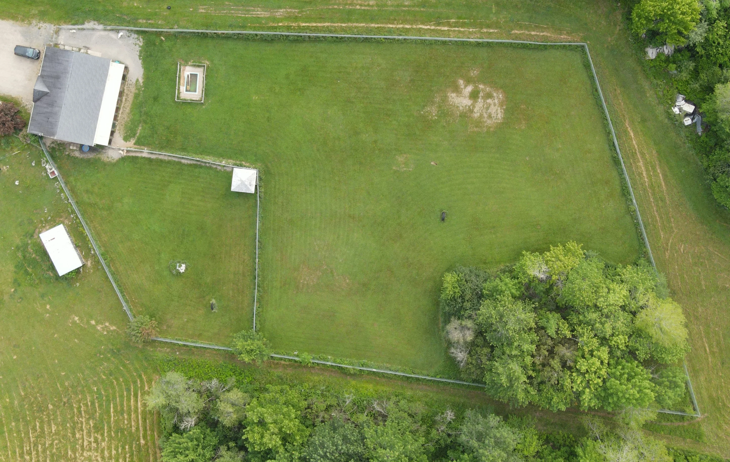 An aerial view of a fenced backyard lawn with some patches, surrounded by trees and neighboring yards, showcasing a house with a driveway and some outdoor structures.