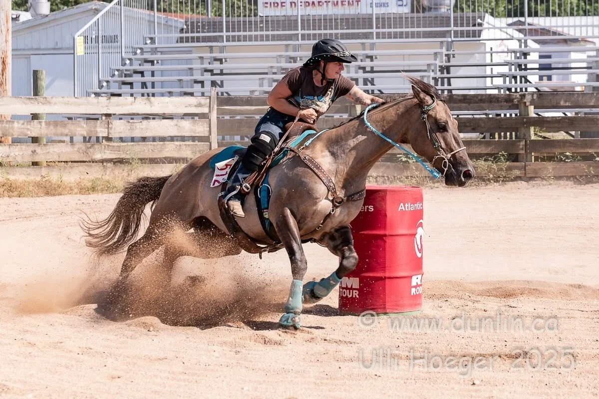 A woman riding a horse around an orange barrel during a rodeo or equestrian event on a sandy arena with wooden fence and bleachers in the background.