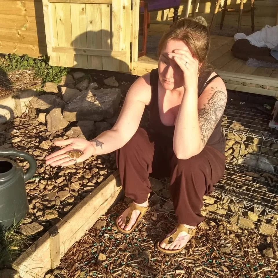 A woman with tattoos sitting on the ground outside, with a butterfly in her left hand, with her right hand on her forehead, looking downward.
