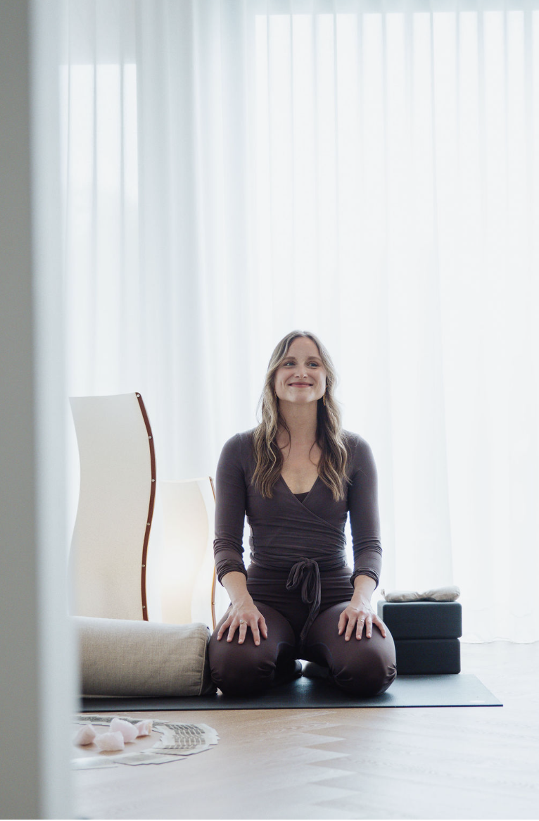 A woman practicing yoga on a mat indoors near sheer white curtains, sitting on her knees with a peaceful expression.
