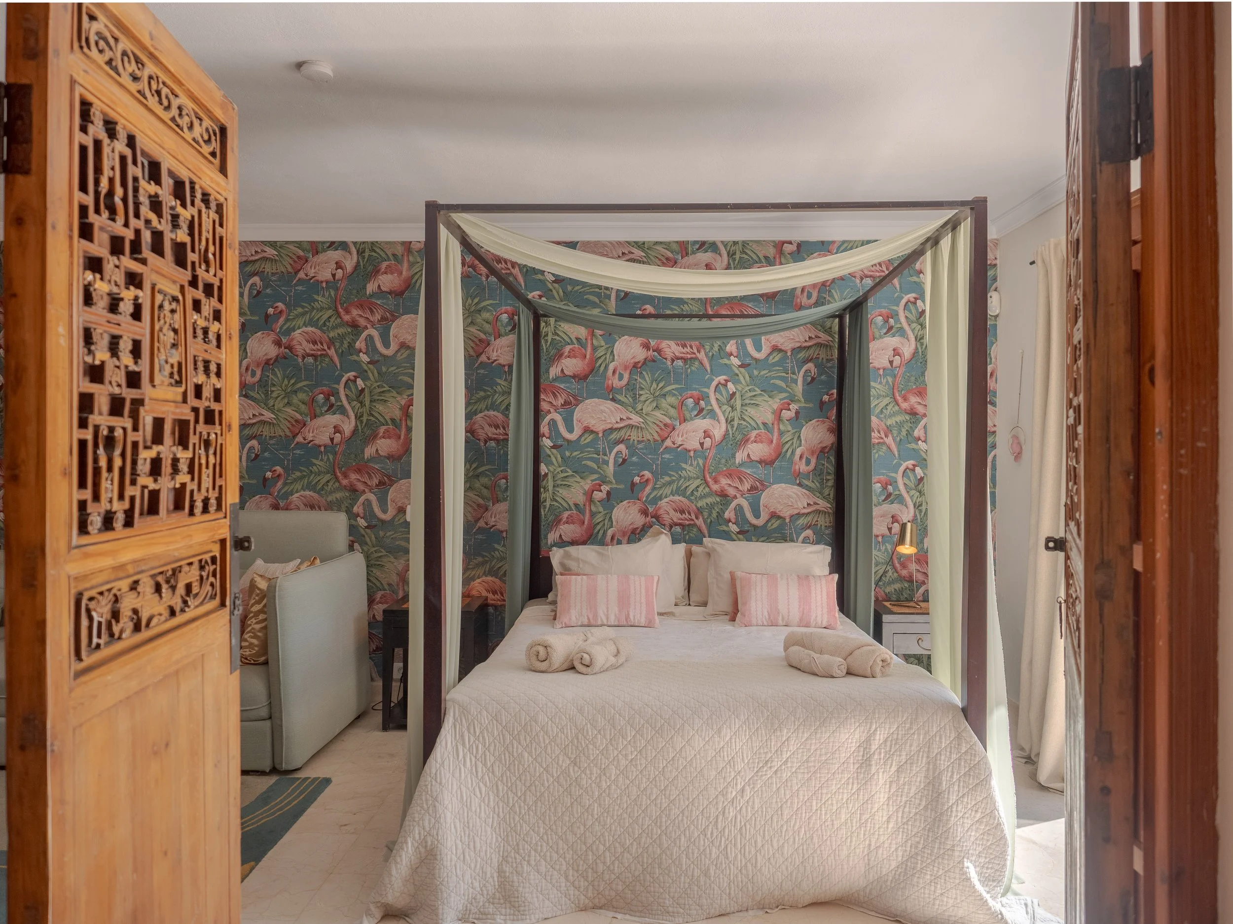 Bedroom with a four-poster bed with beige quilt and pink pillows, flamingo wallpaper, and a towel set on the bed.