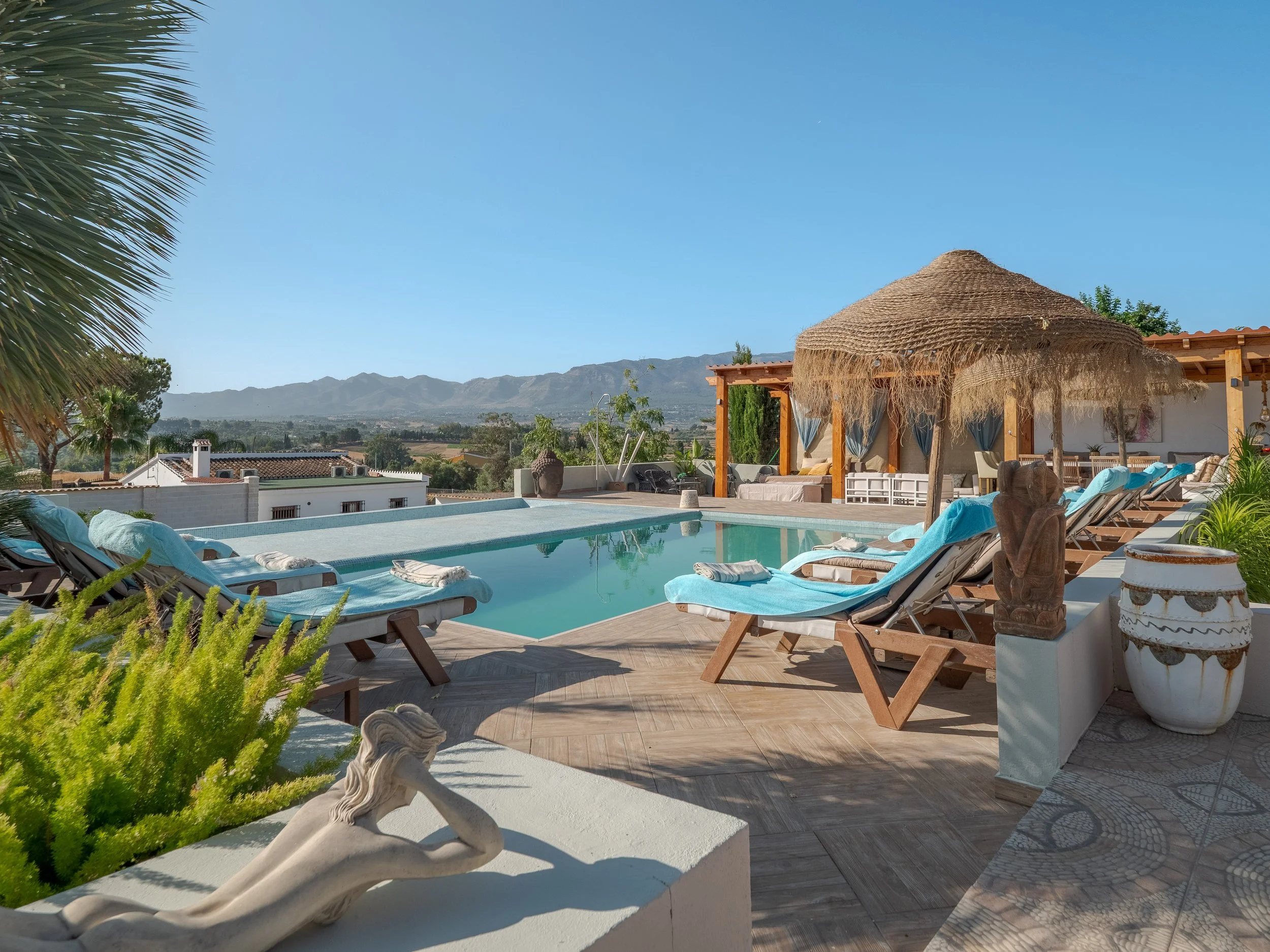 An outdoor pool area with lounge chairs, a thatched umbrella, green plants, a white statue of a reclining figure, and a scenic mountain view under a blue sky.