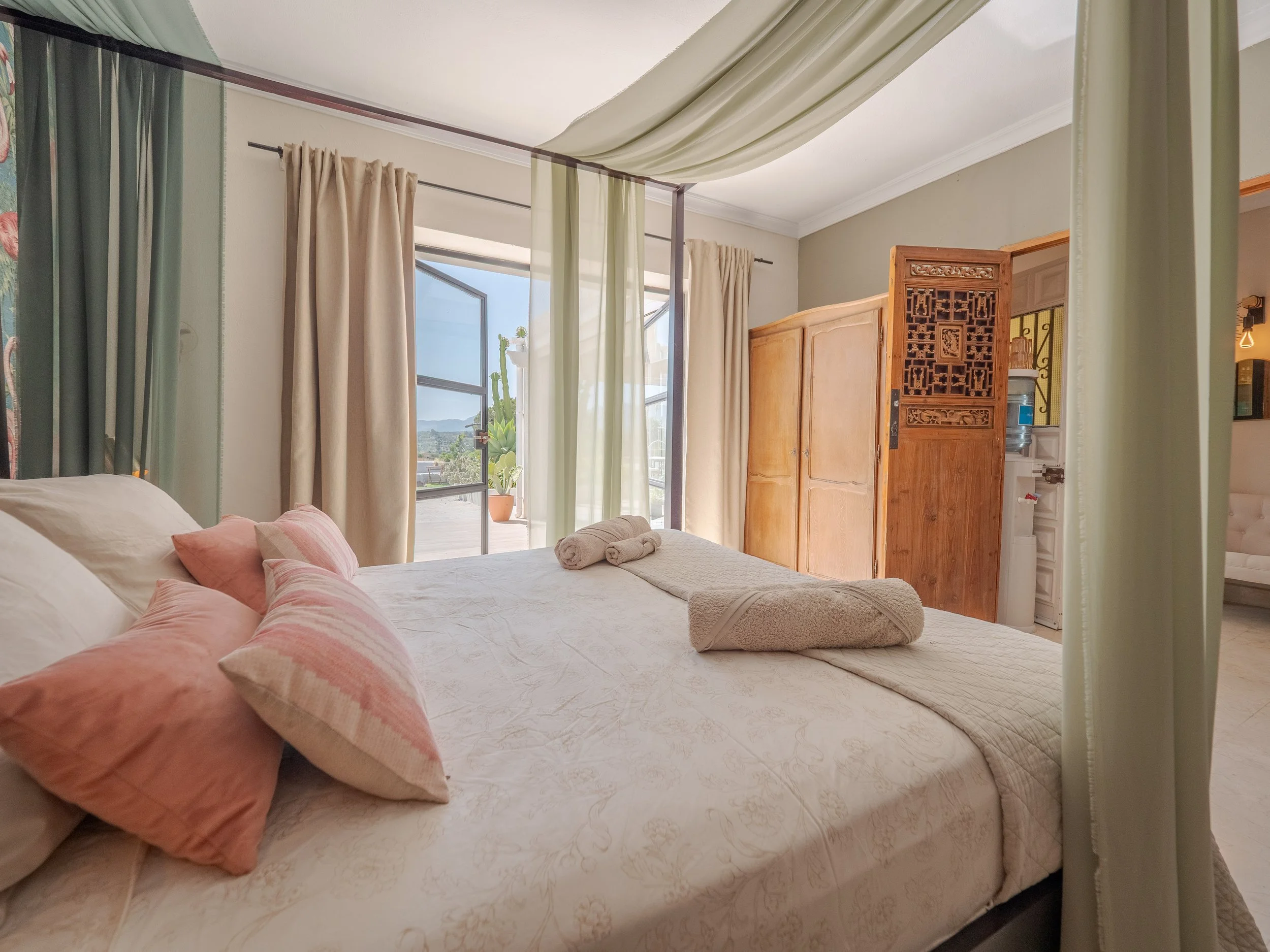 A bedroom with beige curtains, a large bed with pink pillows, a wooden wardrobe, and a sliding glass door leading to a balcony with a potted cactus plant outside.