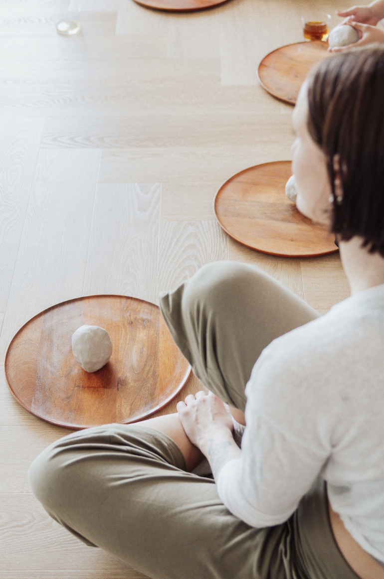A person sitting cross-legged on the floor with wooden plates in front of them. There are small objects or food items on each plate, and they appear to be participating in an activity involving the items on the plates.