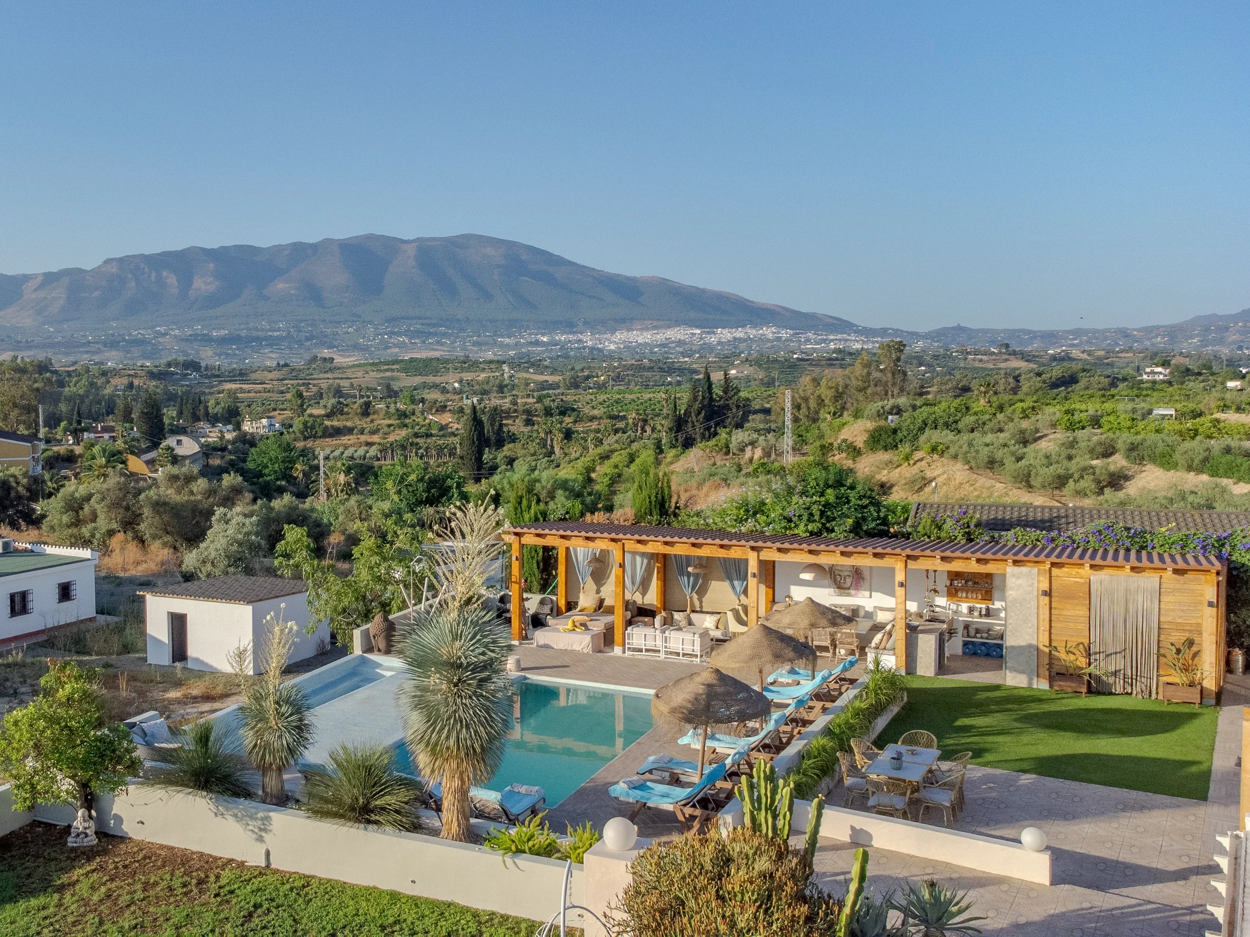 A backyard with a swimming pool, lounge chairs, and a shaded outdoor seating area. There are trees, plants, and mountains in the background under a clear blue sky.
