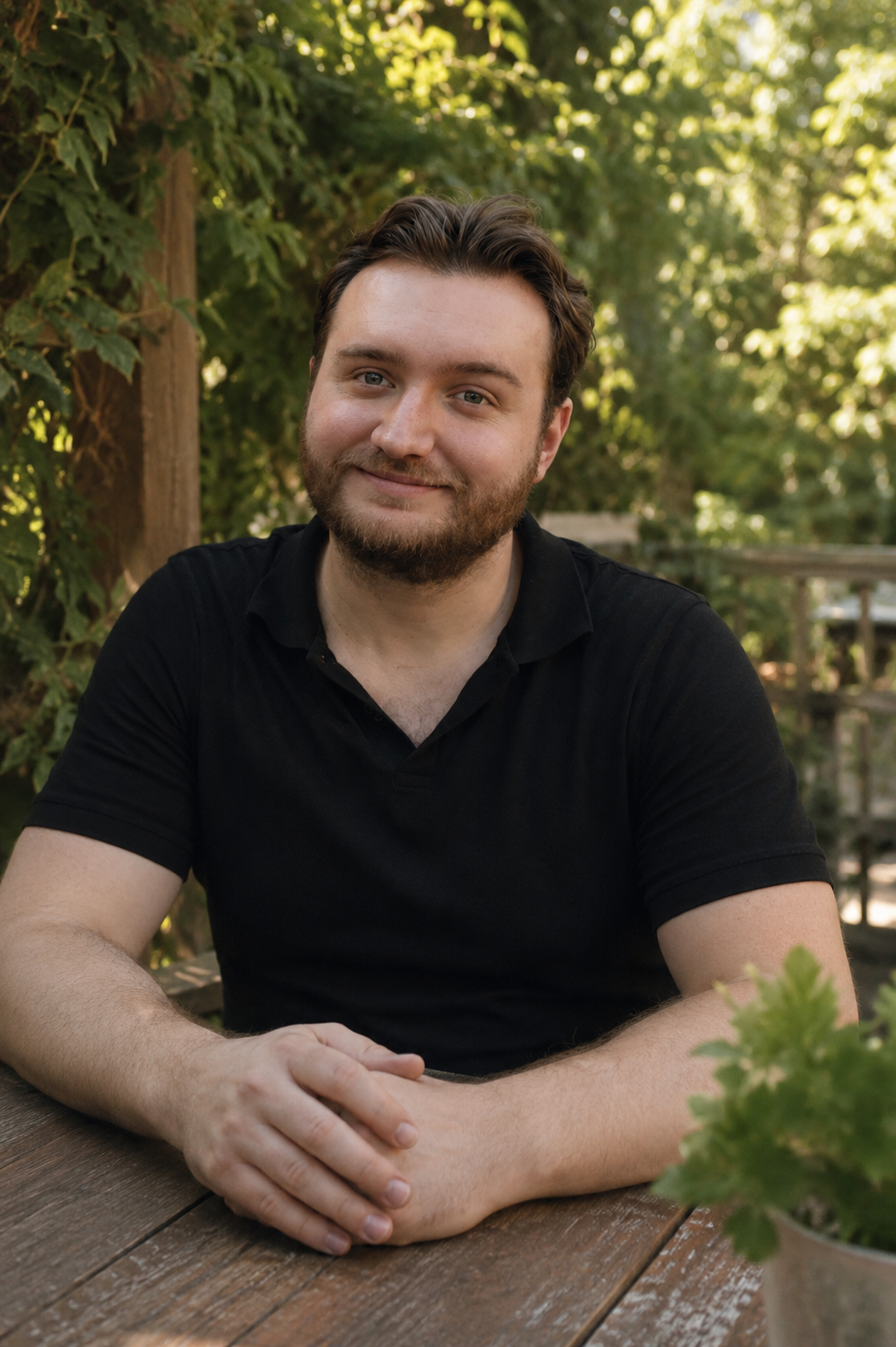 Un homme souriant avec une barbe, assis à une table en bois dans un environnement extérieur avec des arbres verts en arrière-plan, portant un t-shirt noir.