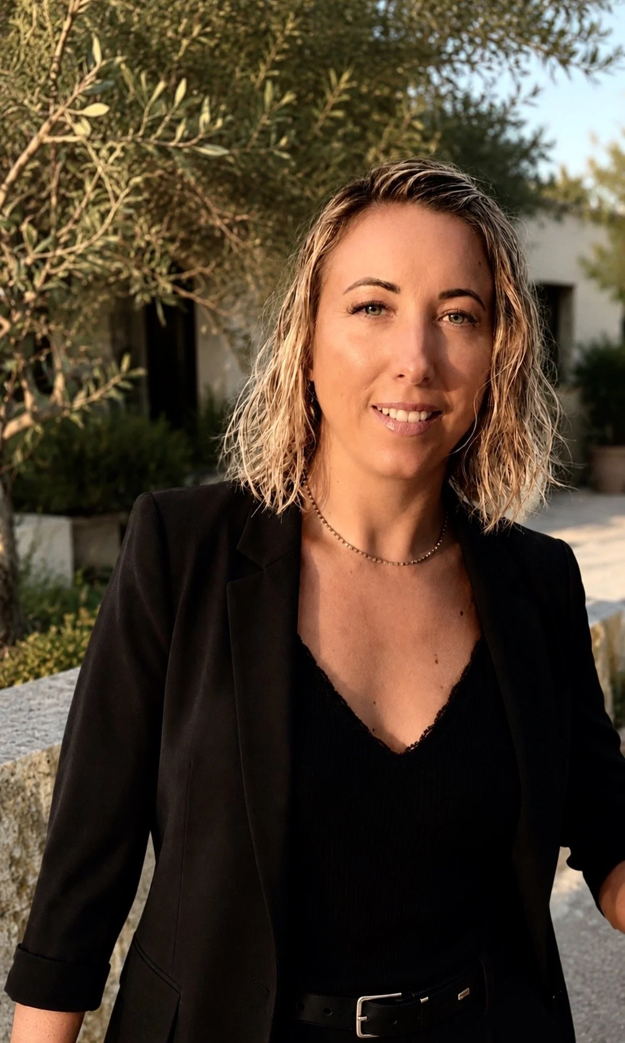 Portrait d'une femme blonde avec des cheveux ondulés, portant un blazer noir et une chaîne, en extérieur avec un arbre en arrière-plan lors du coucher de soleil.