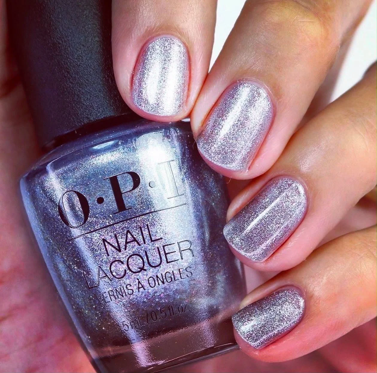 D'ongles peints avec du vernis à ongles argenté irisé, tenu par une main. Le flacon de vernis à ongles O.P.I est visible.