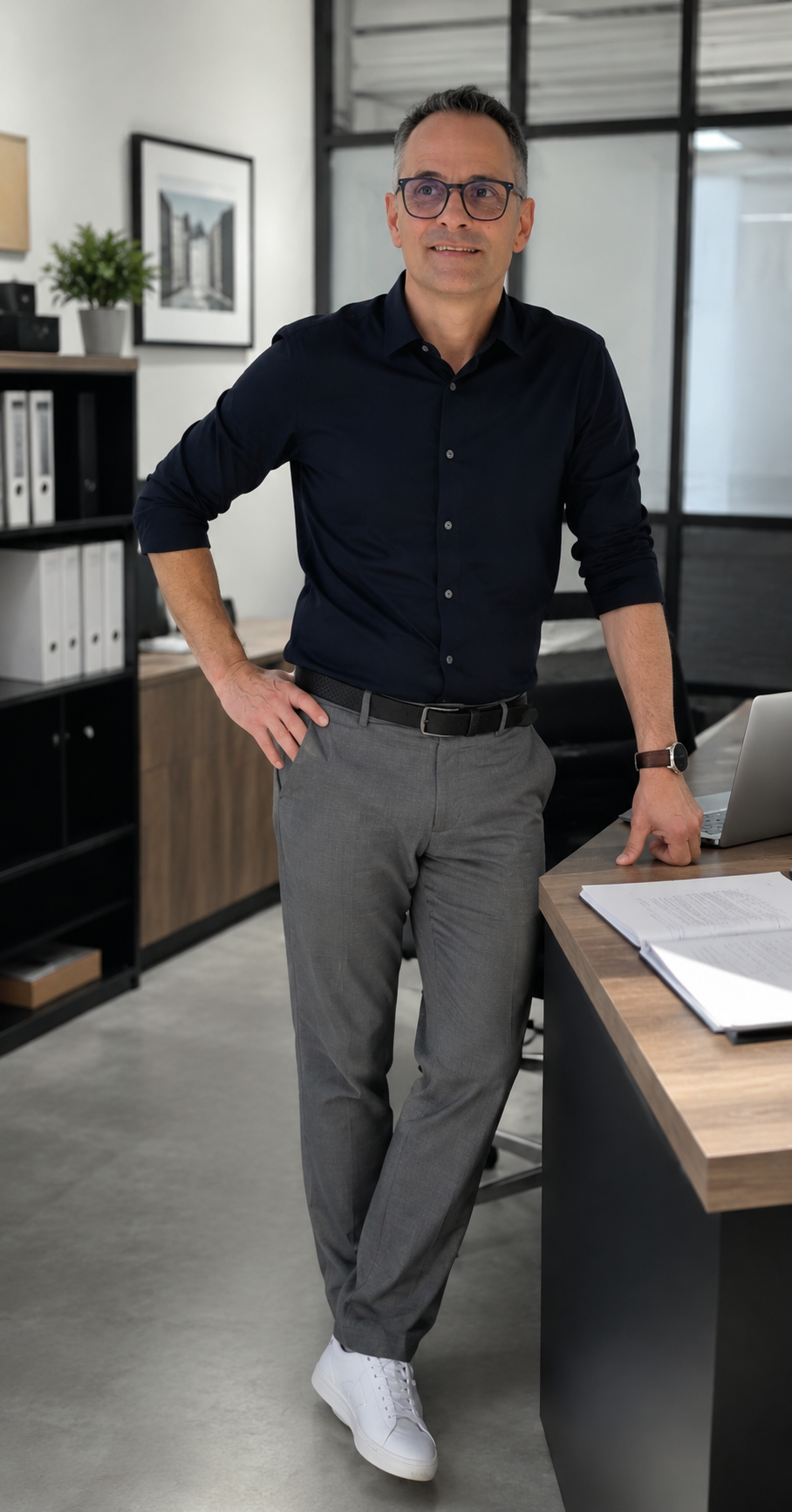 Un homme d'affaires debout dans un bureau moderne avec une main sur la table, portant une chemise noire, un pantalon gris, des lunettes, une montre et des baskets blanches.
