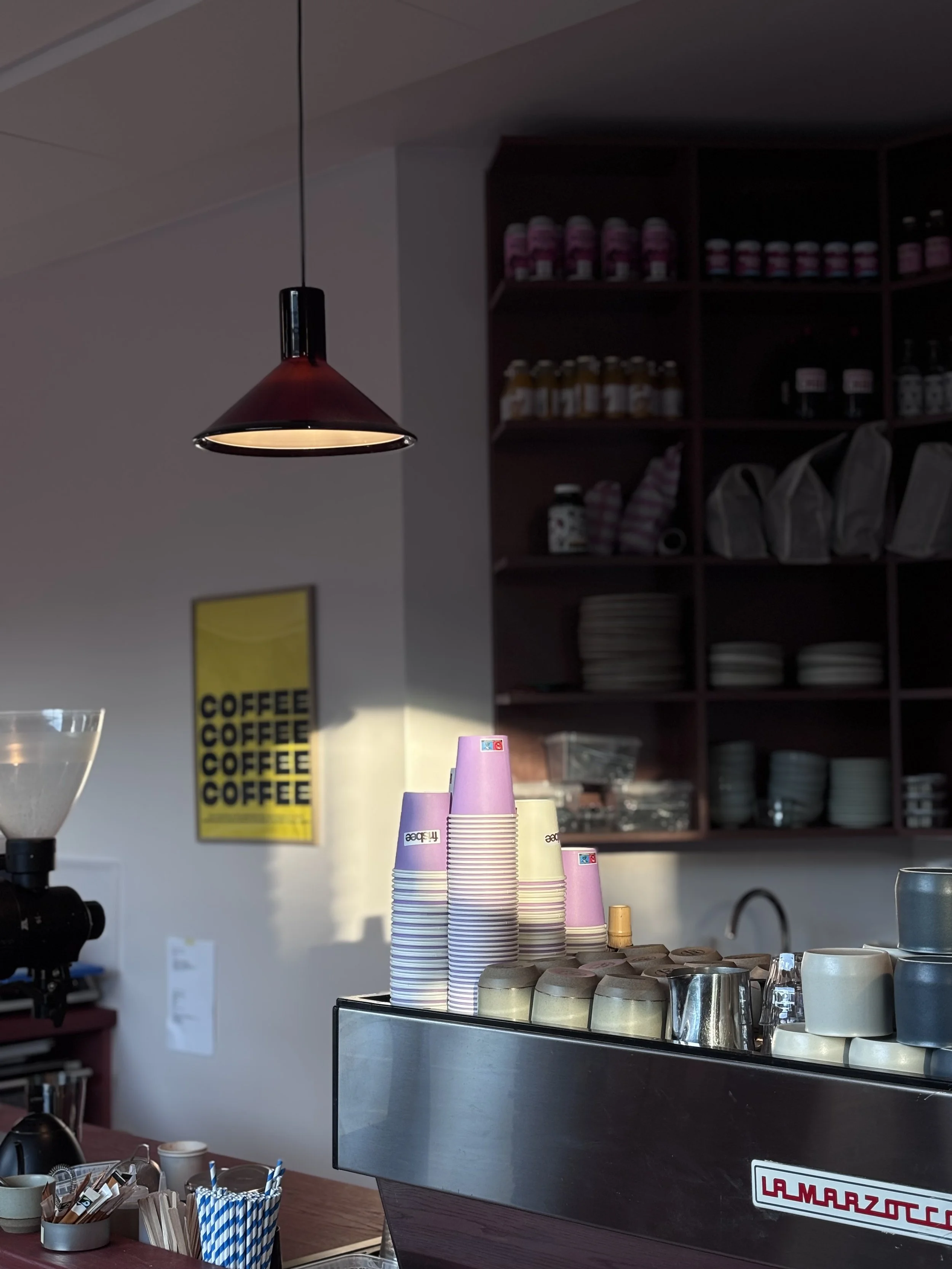 Stacked pink and purple cups on a counter in a coffee shop, with shelves of jars and dishes in the background.