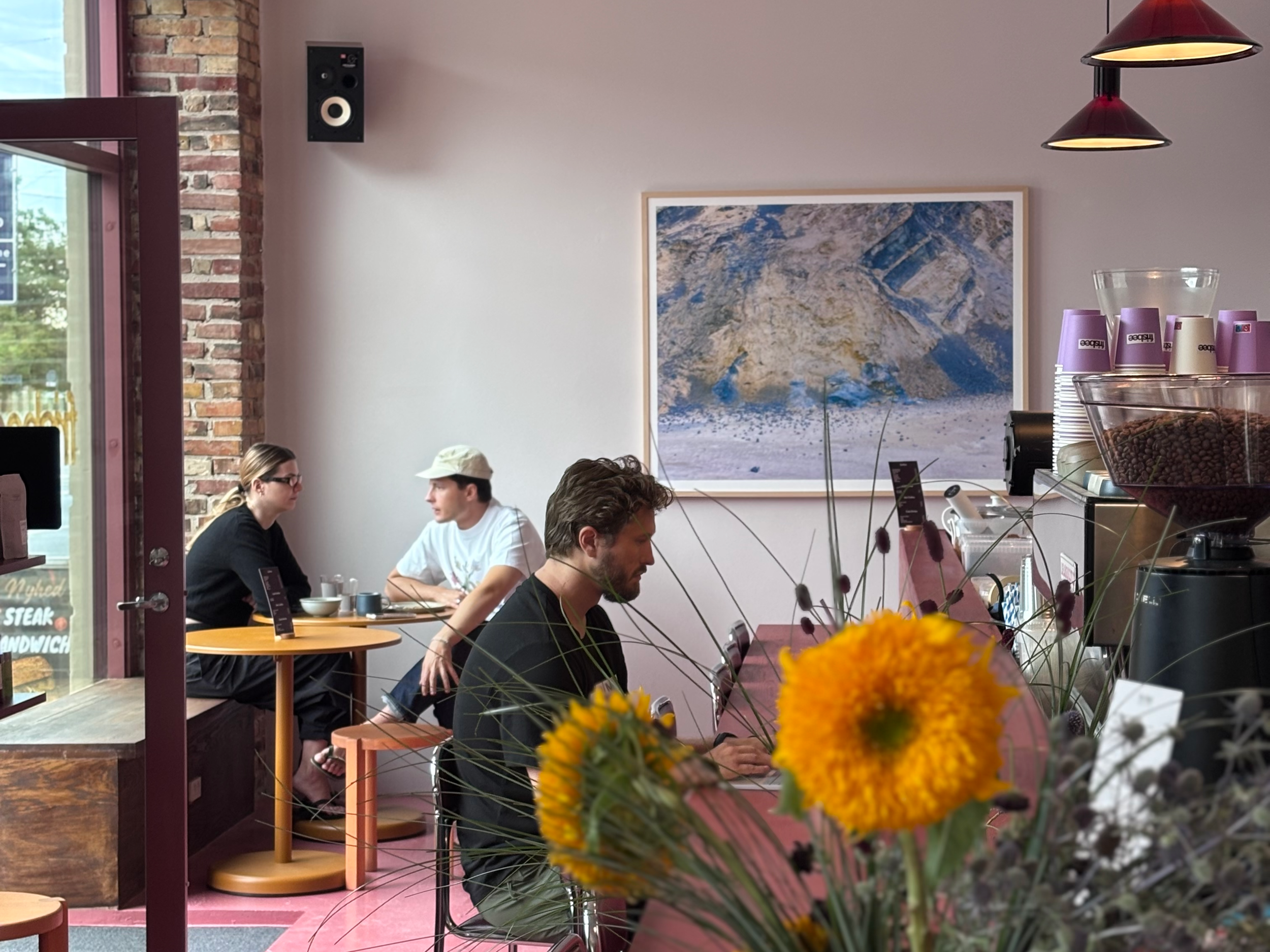 Interior of a cafe with several customers sitting at tables, large mountain artwork on the wall, a flower arrangement with yellow flowers in the foreground, and a display with purple cups on the counter.