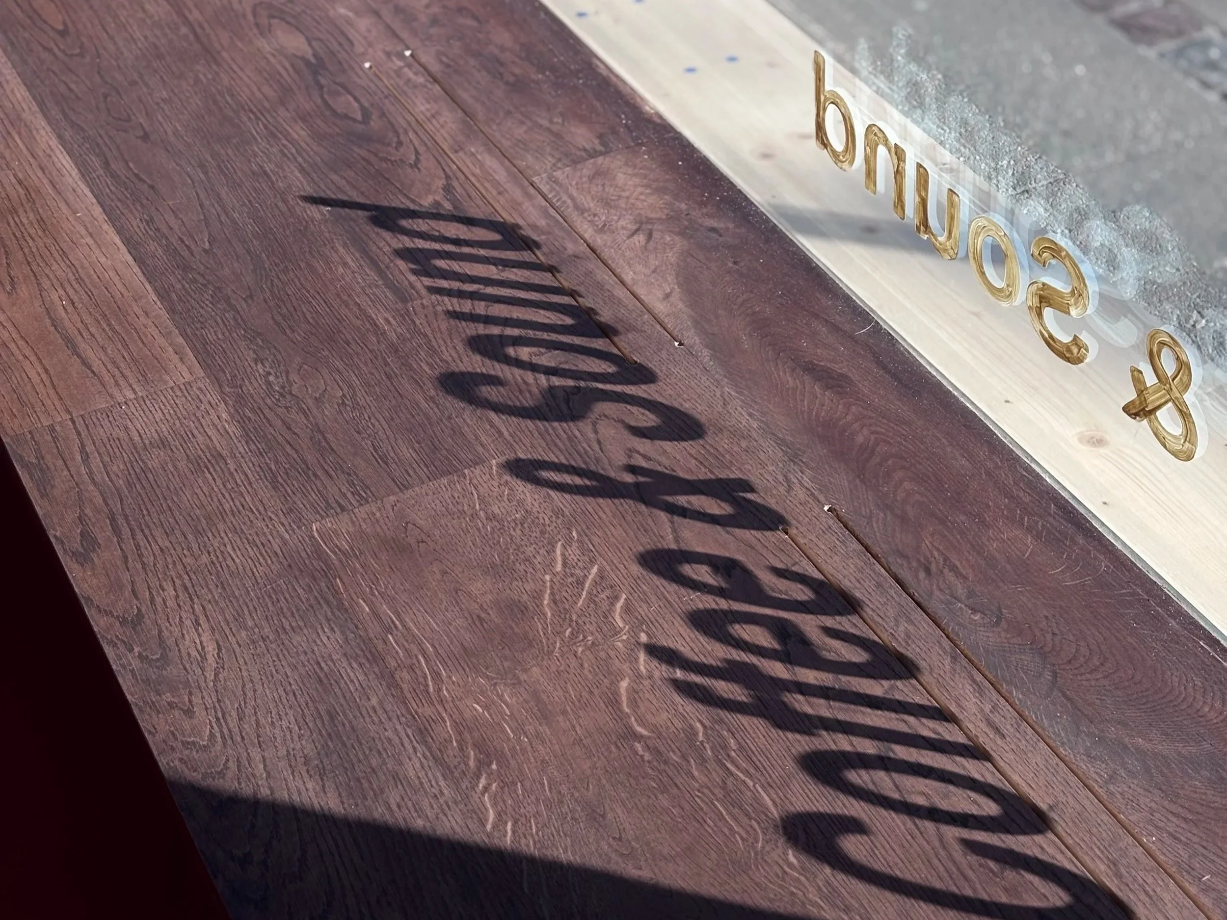 Close-up of a wooden surface with a shadow of decorative lettering cast on it, and a transparent sign with gold-embossed letters leaning against a wall.