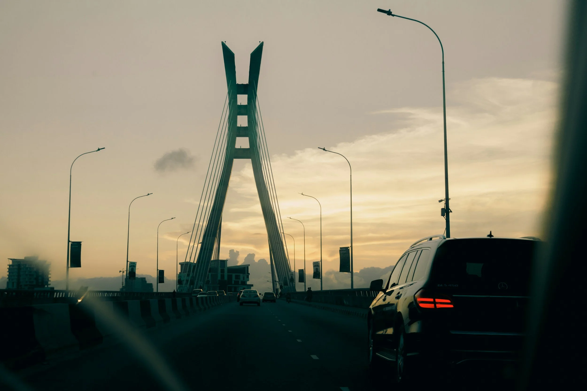 A bridge with a modern, distinctive design viewed from a moving vehicle on a highway at sunset, with streetlights and buildings visible in the background.