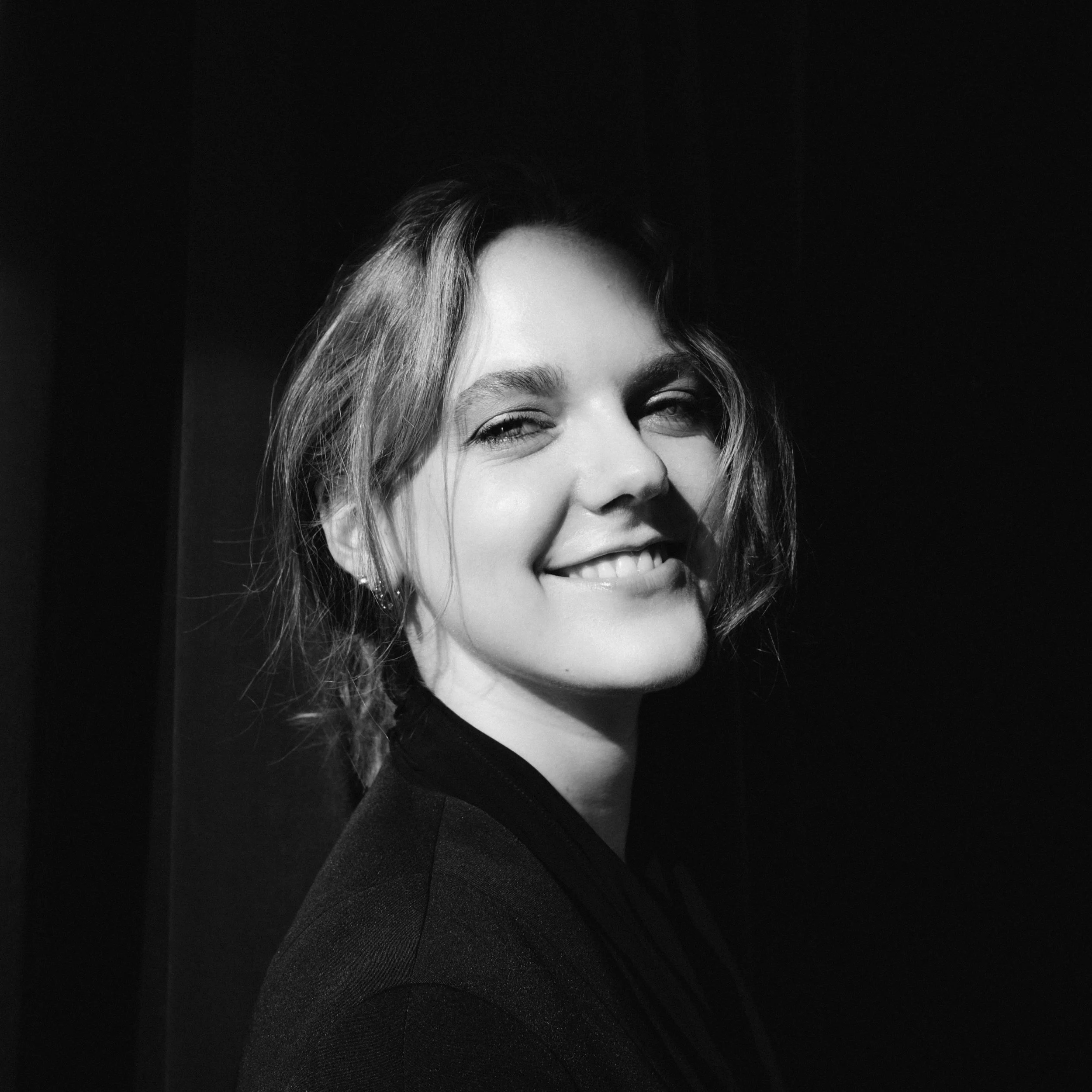 Close-up of a smiling woman with wavy hair, wearing earrings, against a dark background.