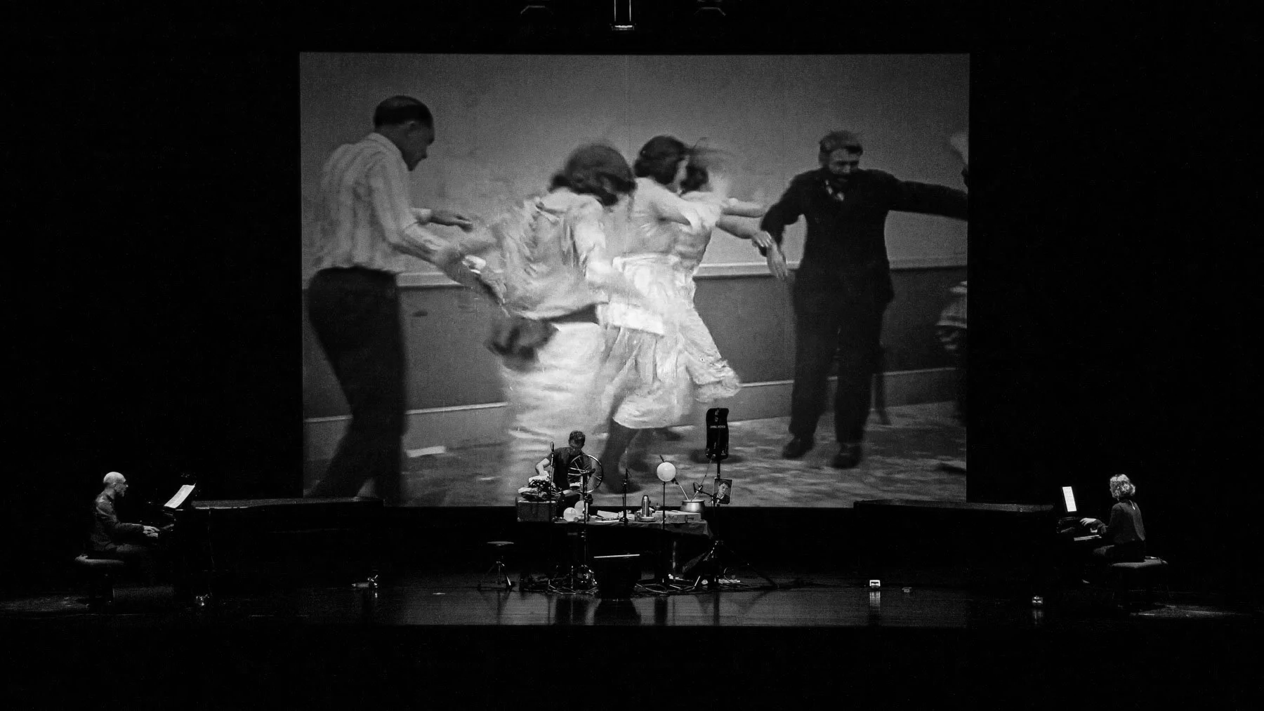 Black-and-white photo displayed on a stage showing women and a man dancing, with musicians playing pianos on either side of the stage.